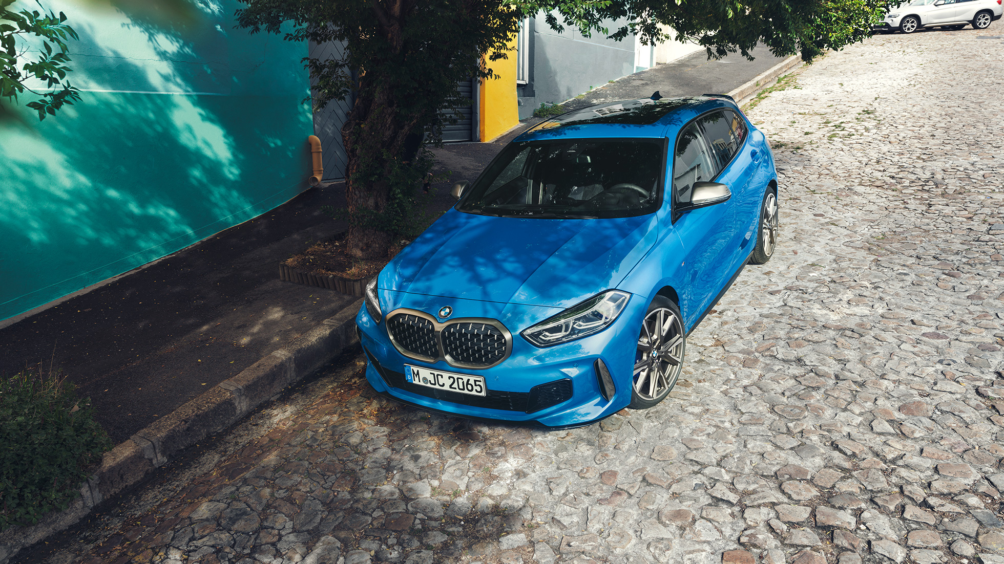 BMW serije 1 na cesti. BMW serije 1 v kovinski barvi Misano Blue, tričetrtinski pogled od spredaj, parkiranje na cesti iz ptičje perspektive.