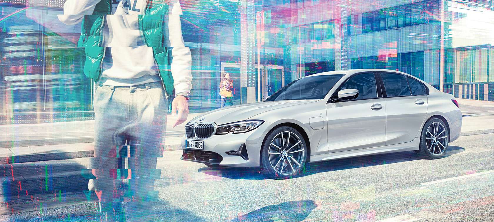 BMW serije 3 Limuzina, stranski posnetek BMW 330e. Tričetrtinski stranski posnetek mirujočega BMW 330e Limuzina pred stekleno stavbo z mimoidočimi.