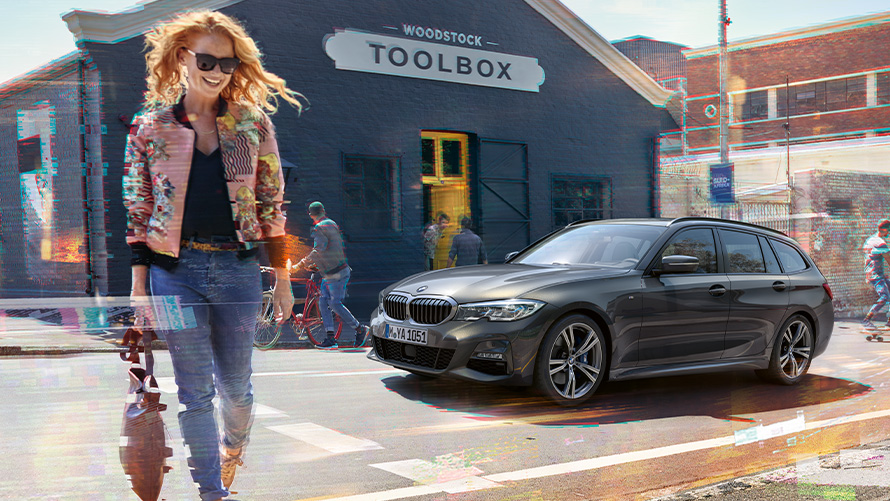 BMW serije 3 Touring (G21), barva tričetrtinski posnetek od spredaj, ženska pred orodjarno. Ženska prečka cesto pred BMW serije 3 Touring (G21) nasproti orodjarne.