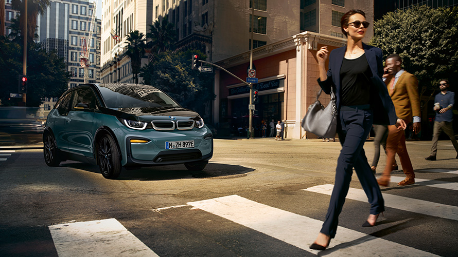 BMW i3, I01, tričetrtinski pogled od spredaj z žensko, ki prečka prehod za pešce BMW i3, I01, 2018, Blue Ridge Mountain s poudarki v kovinski barvi Frozen Grey, tričetrtinski pogled od spredaj z žensko, ki prečka prehod za pešce