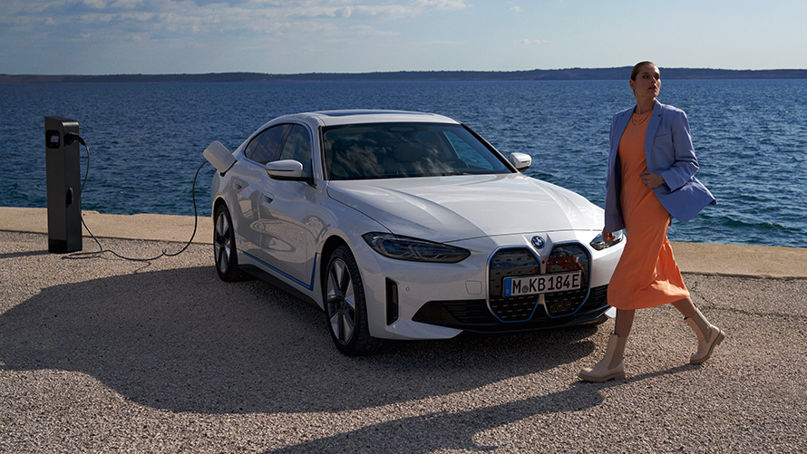 BMW i4 eDrive40, G26, 2021, tričetrtinski pogled od spredaj na polnilno postajo Ženska stoji pred BMW i4 eDrive40, G26, 2021, barva Mineral White, tričetrtinski pogled od spredaj na polnilno postajo pred jezerom