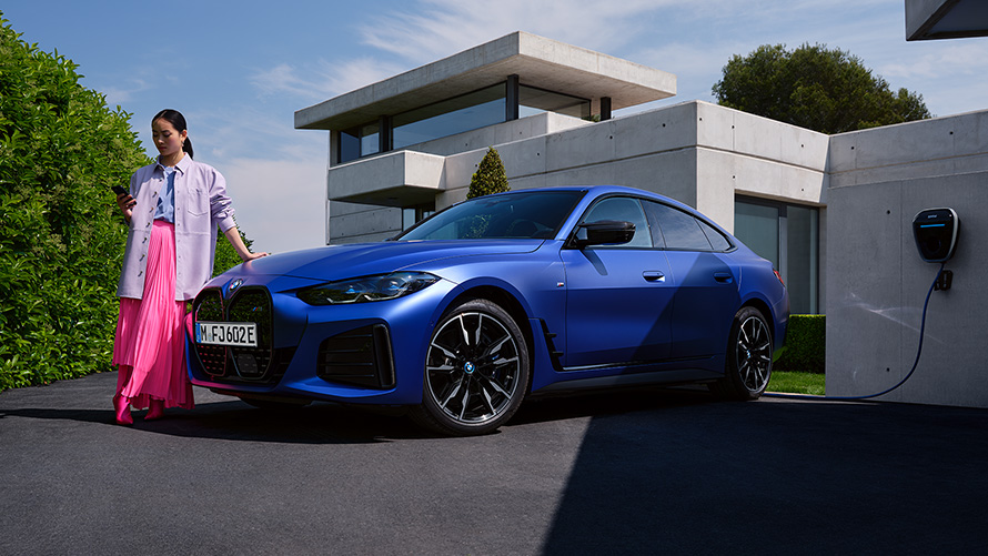 BMW i4 M50, G26, 2021, lakiranje BMW Individual v kovinski barvi Frozen Portimao Blue, tričetrtinski pogled od spredaj med mirovanjem z žensko pred hišo