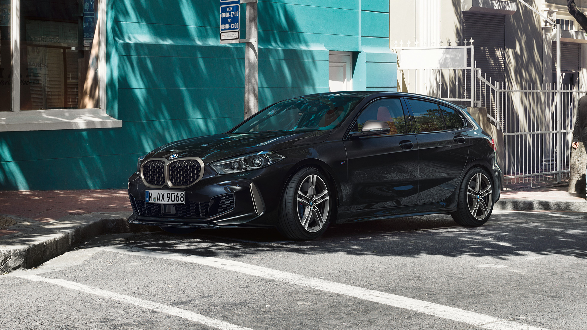 BMW M135i xDrive, tričetrtinski stranski pogled. Popolnoma novi BMW M135i xDrive v kovinski barvi Black Sapphire, tričetrtinski stranski pogled na parkirišče.