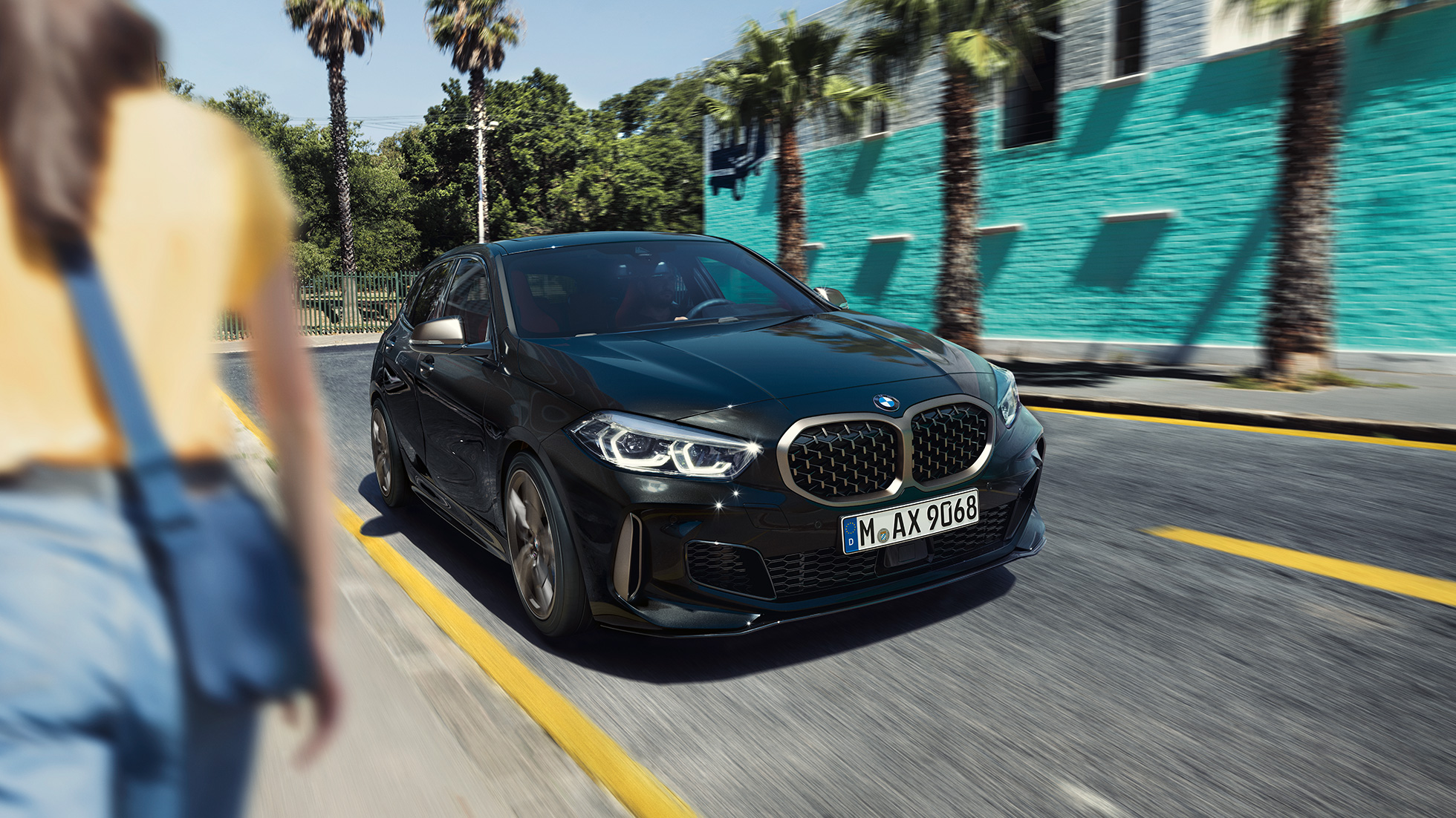 BMW M135i xDrive, tričetrtinski pogled od spredaj. BMW M135i xDrive v kovinski barvi Black Sapphire, tričetrtinski pogled od spredaj med vožnjo z žensko v ospredju.