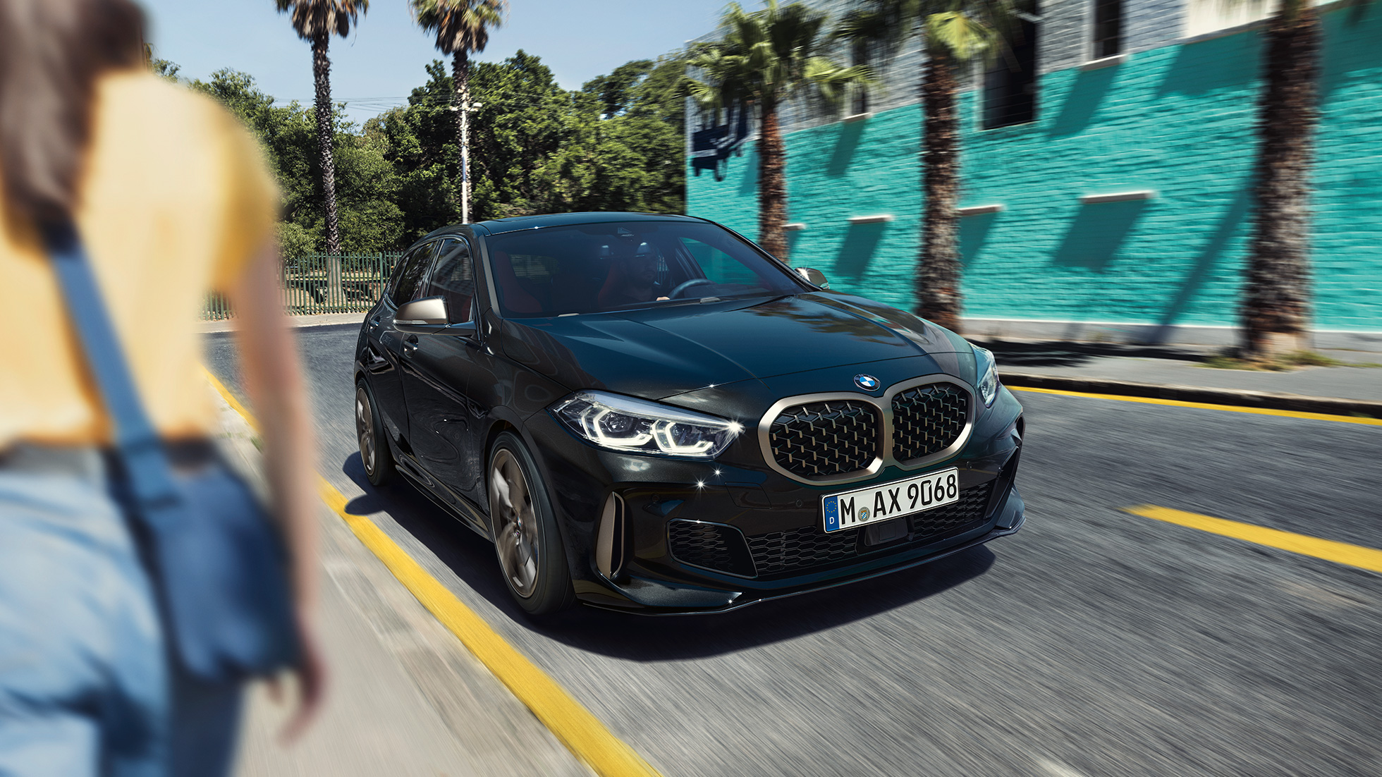 BMW M135i xDrive, tričetrtinski pogled od spredaj. BMW M135i xDrive v kovinski barvi Black Sapphire, tričetrtinski pogled od spredaj med vožnjo z žensko v ospredju.