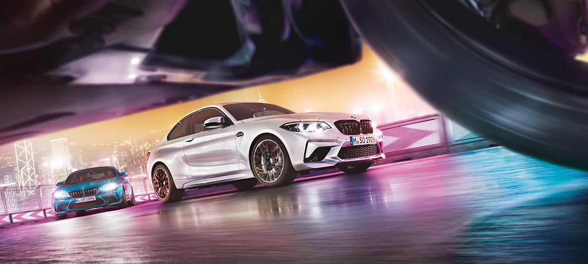 BMW M2 Competition na odsevnem asfaltu Dva BMW M2 Competition na stezi pred nočno kuliso velemesta z vidika ceste.