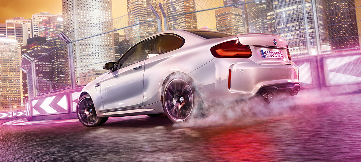 BMW M2 Competition dodaja plin Bel BMW M2 Competition drsi bočno na stezi pred nočnim mestnim obzorjem.