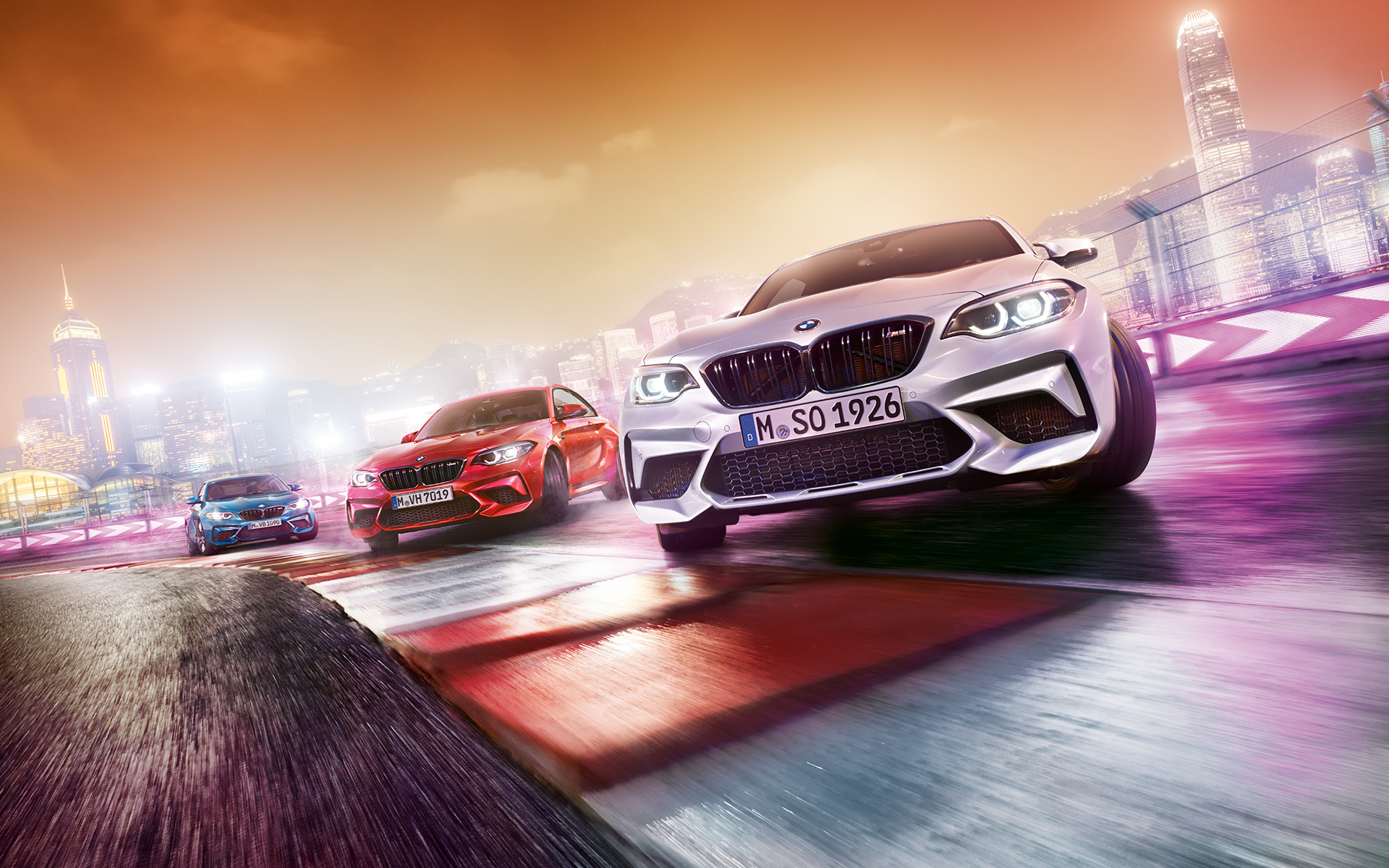 BMW M2 Competition v akciji Trije BMW M2 Competition drsijo bočno na stezi pred nočno kuliso velemesta.