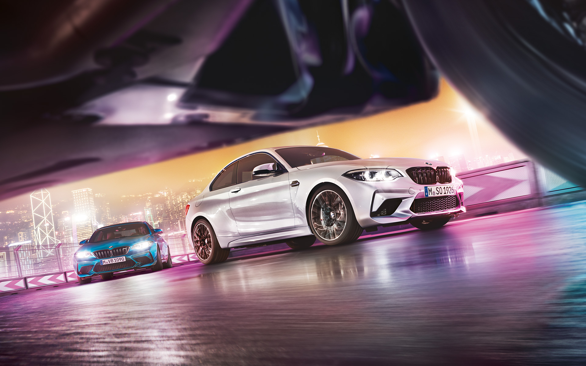 BMW M2 Competition na odsevnem asfaltu Dva BMW M2 Competition na stezi pred nočno kuliso velemesta z vidika ceste.