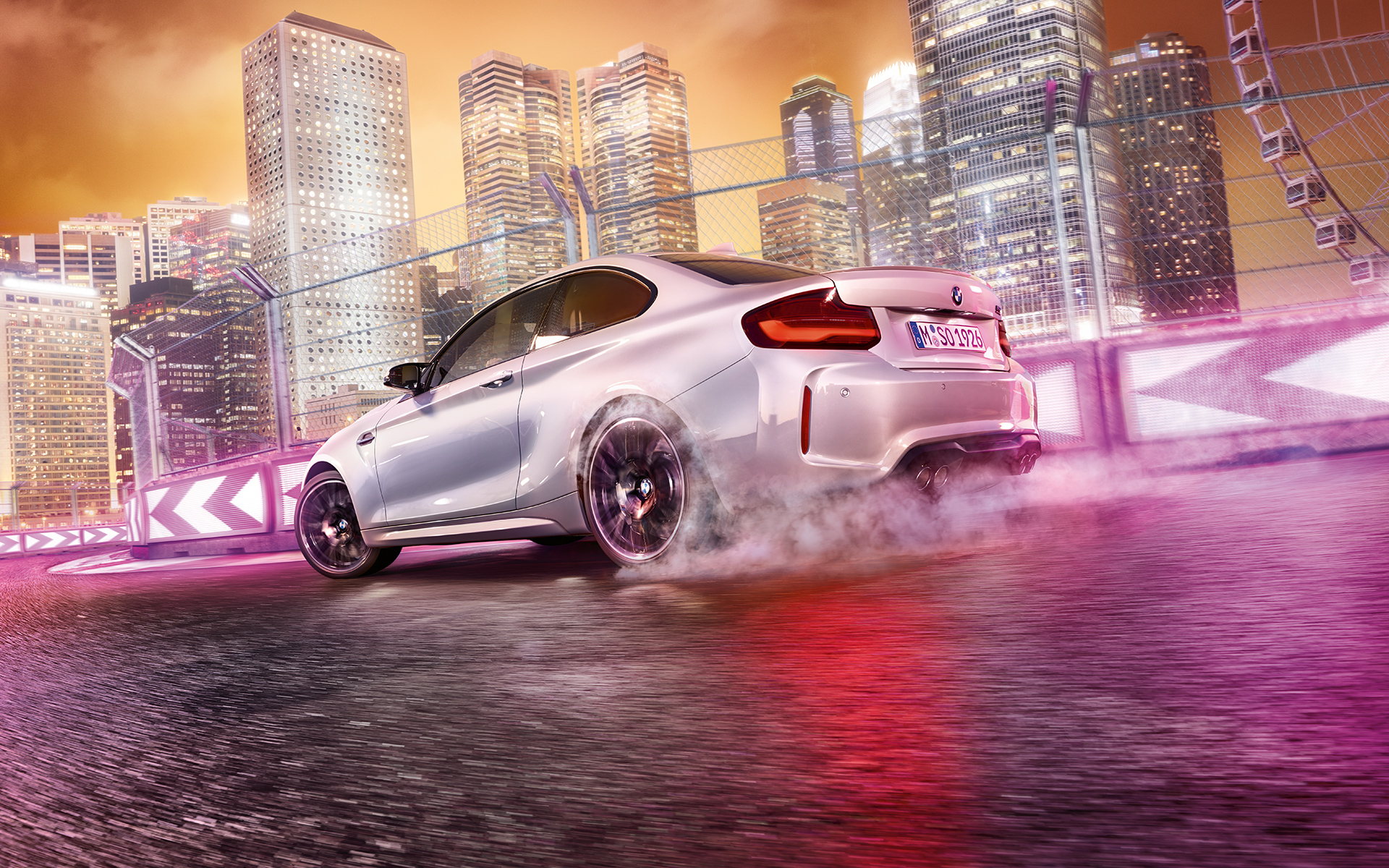 BMW M2 Competition dodaja plin Bel BMW M2 Competition drsi bočno na stezi pred nočnim mestnim obzorjem.