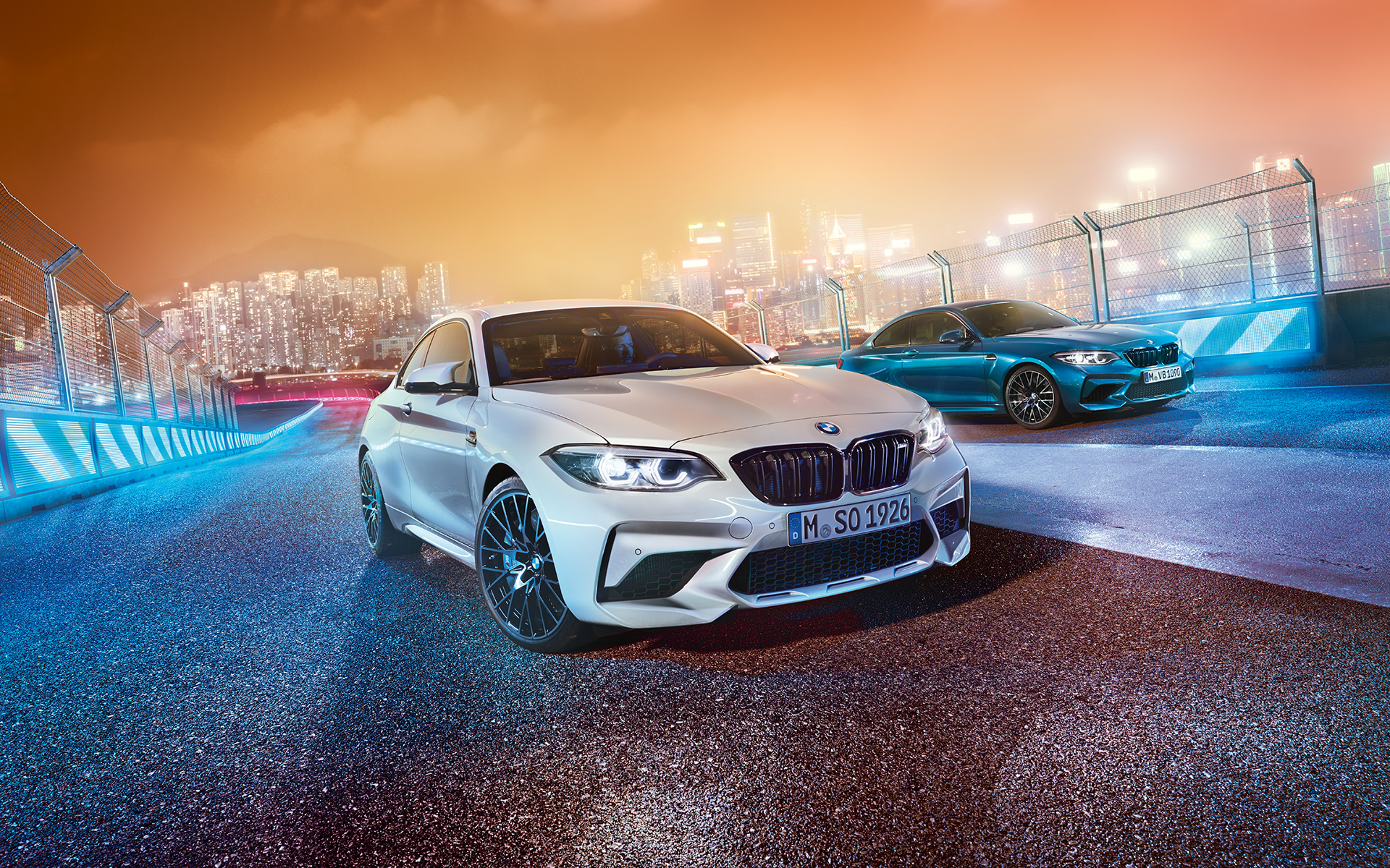 Dva BMW M2 Competition na stezi Bel in moder BMW M2 Competition stojita poševno pred nočno kuliso velemesta v pogledu od spredaj.