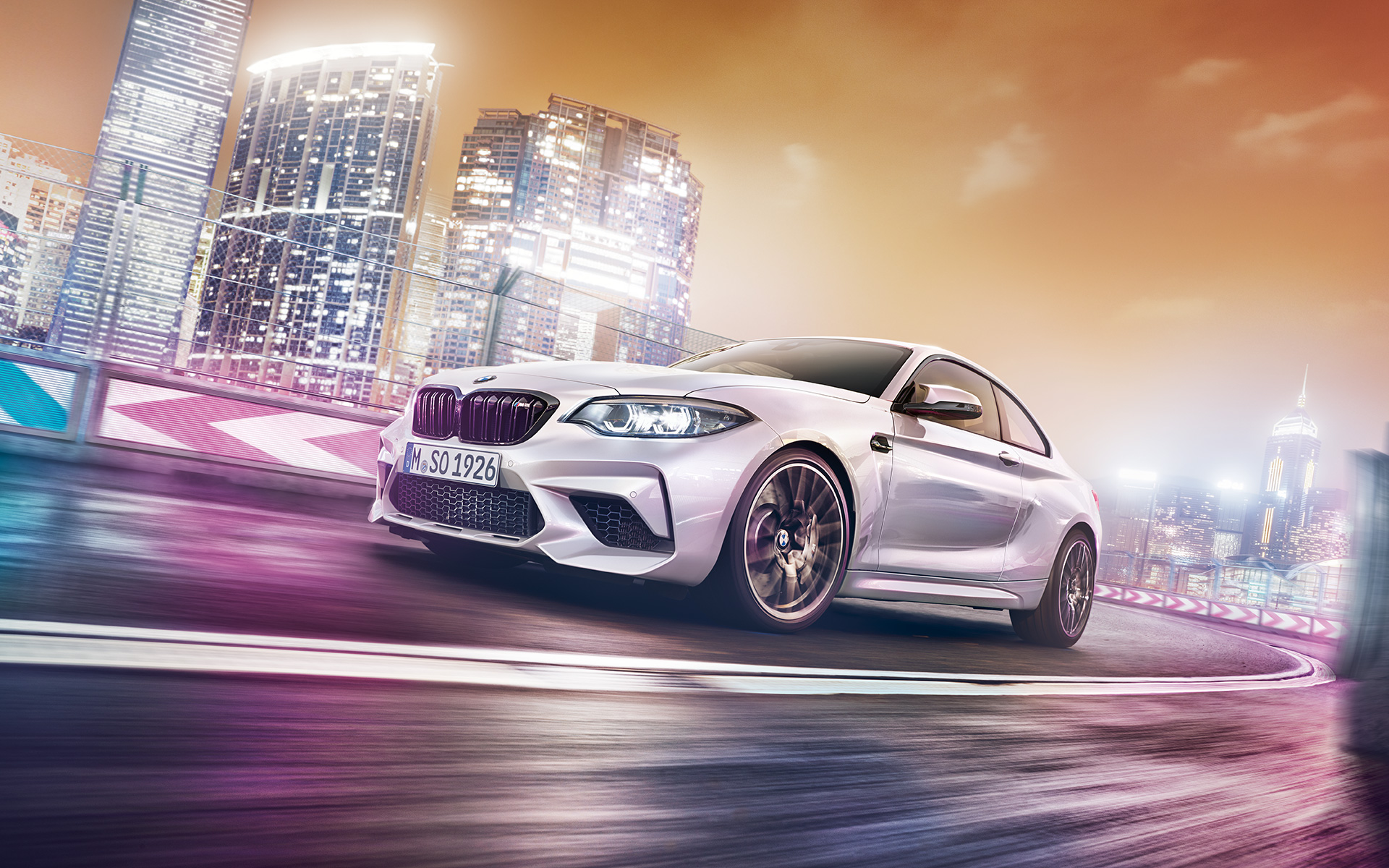 Bel BMW M2 Competition z nebotičnikom Bel BMW M2 Competition vozi na stezi pred nočnim mestnim obzorjem.