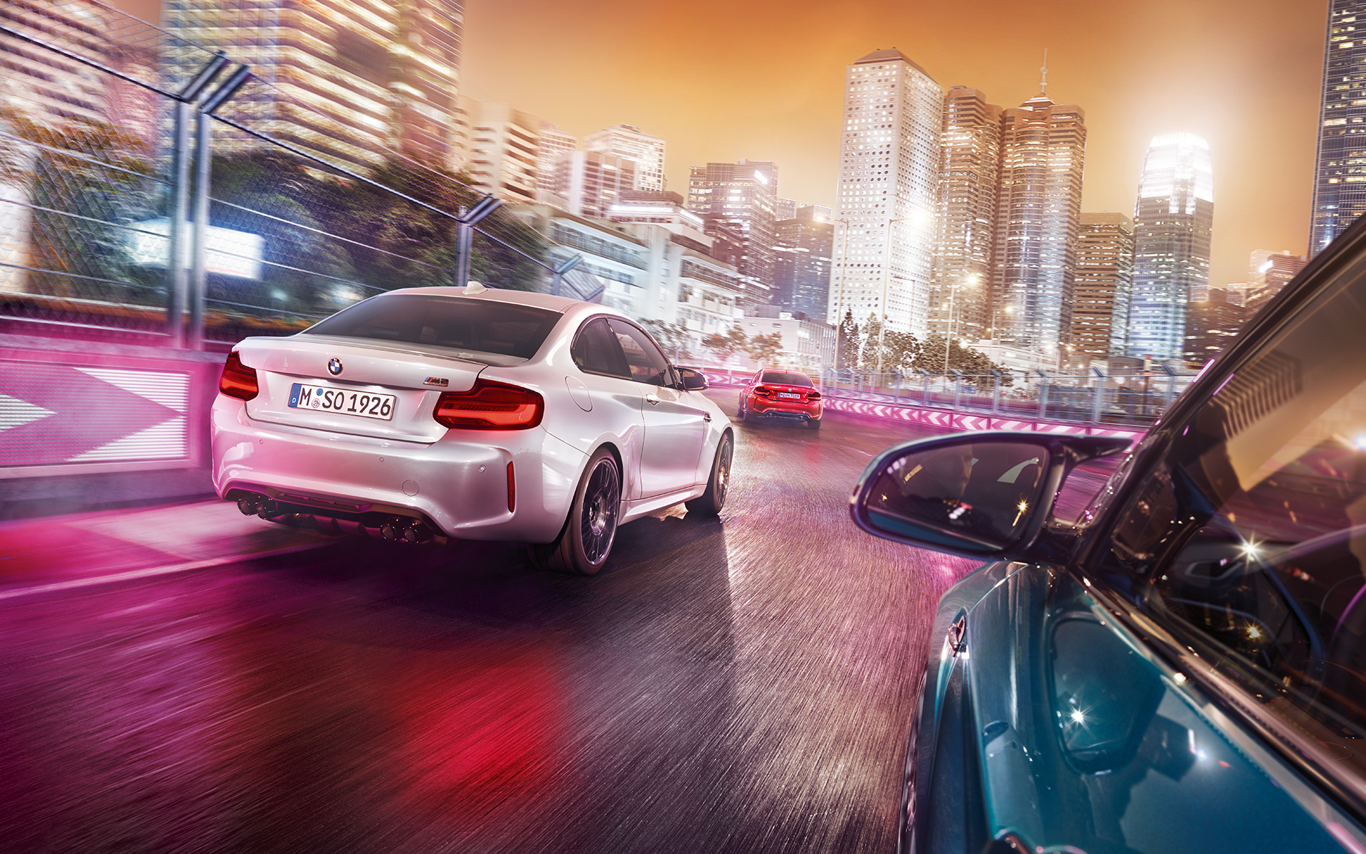 BMW M2 Competition v pogledu od zadaj Trije BMW M2 Competition vozijo na stezi pred osvetljenim nebotičnikom v pogledu od zadaj.