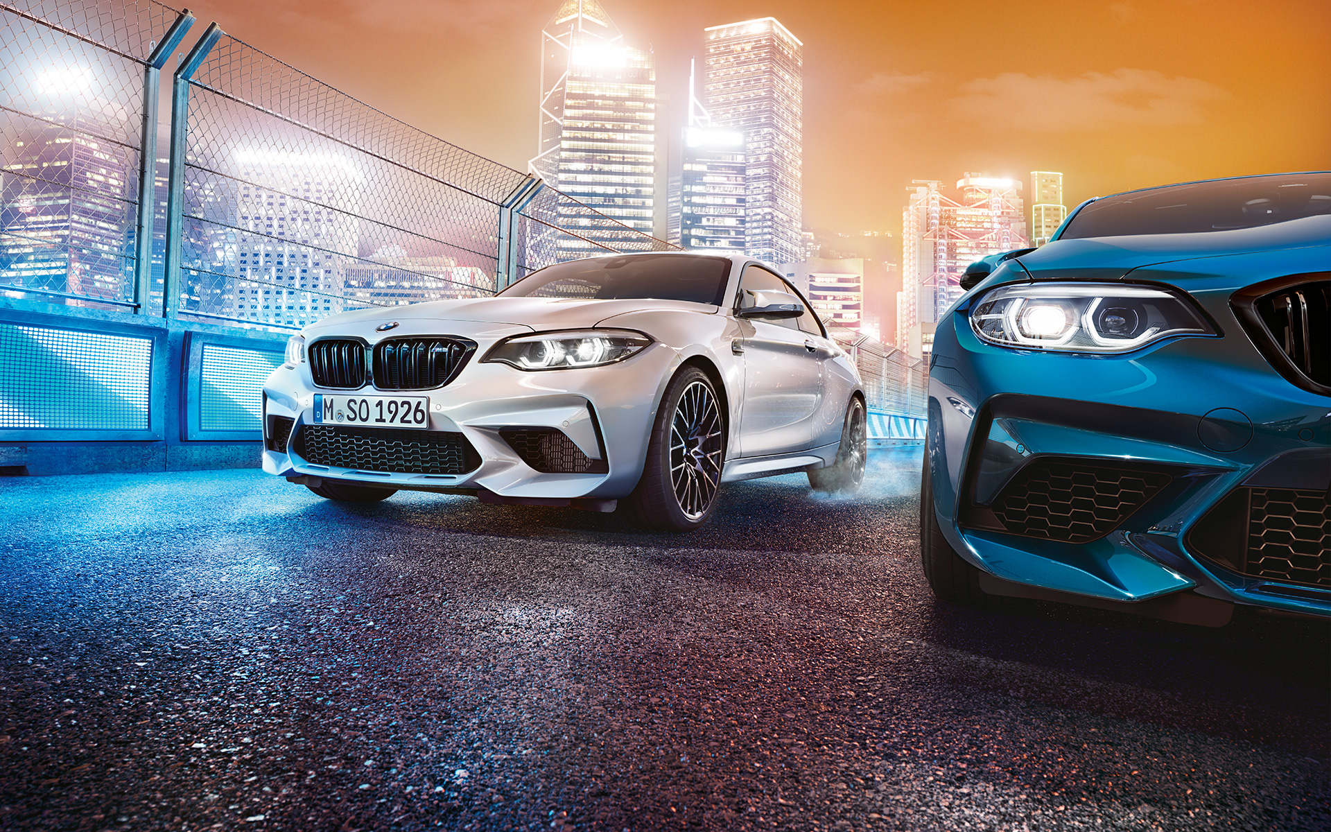 BMW M2 Competition v pogledu od spredaj Bel in moder BMW M2 Competition stojita pred nočno kuliso velemesta v pogledu od spredaj.