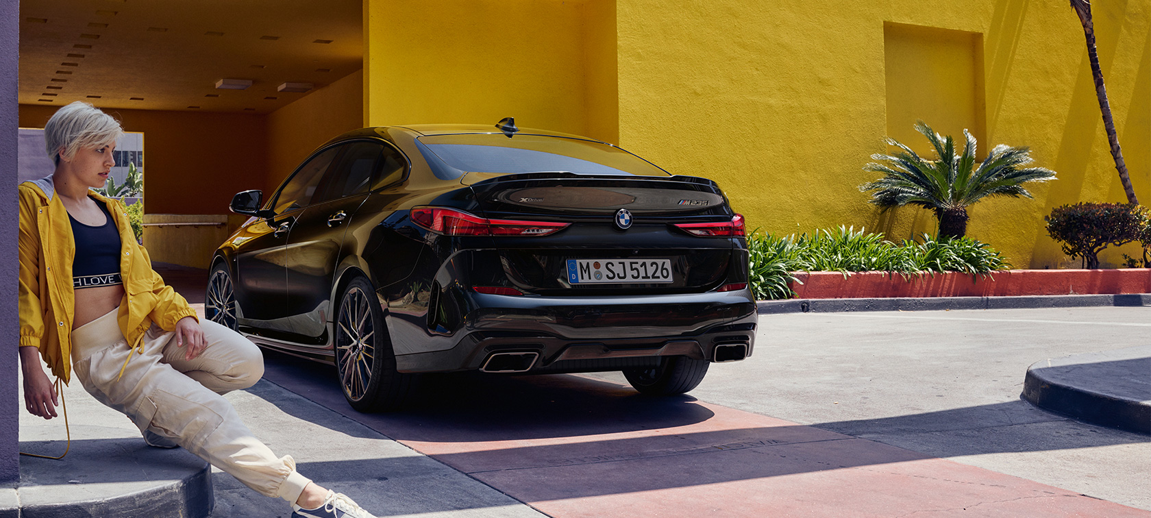 BMW M235i xDrive Gran Coupé, tričetrtinski pogled od zadaj BMW M235i xDrive Gran Coupé v kovinski barvi Black Sapphire F44, tričetrtinski pogled od zadaj z žensko v ospredju