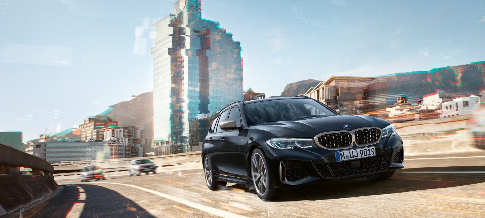 Tričetrtinski posnetek od spredaj med vožnjo pred mestnim ozadjem BMW M340i xDrive Touring, tričetrtinski posnetek od spredaj med vožnjo pred mestnim ozadjem