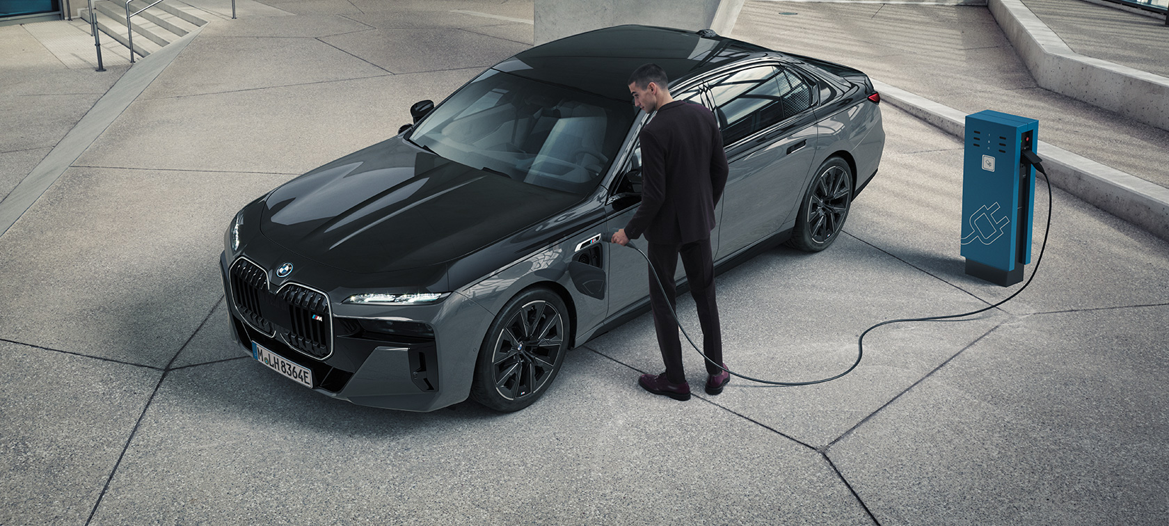 BMW M760e xDrive Limuzina, G70, paket M Performance BMW M760e xDrive Limuzina, G70, paket M Performance, dvobarvno lakiranje BMW Individual v kovinski barvi Black Sapphire, lakiranje BMW Individual v kovinski barvi Dravite Grey, tričetrtinski pogled od spredaj, moški s polnilnim kablom poleg polnilne postaje
