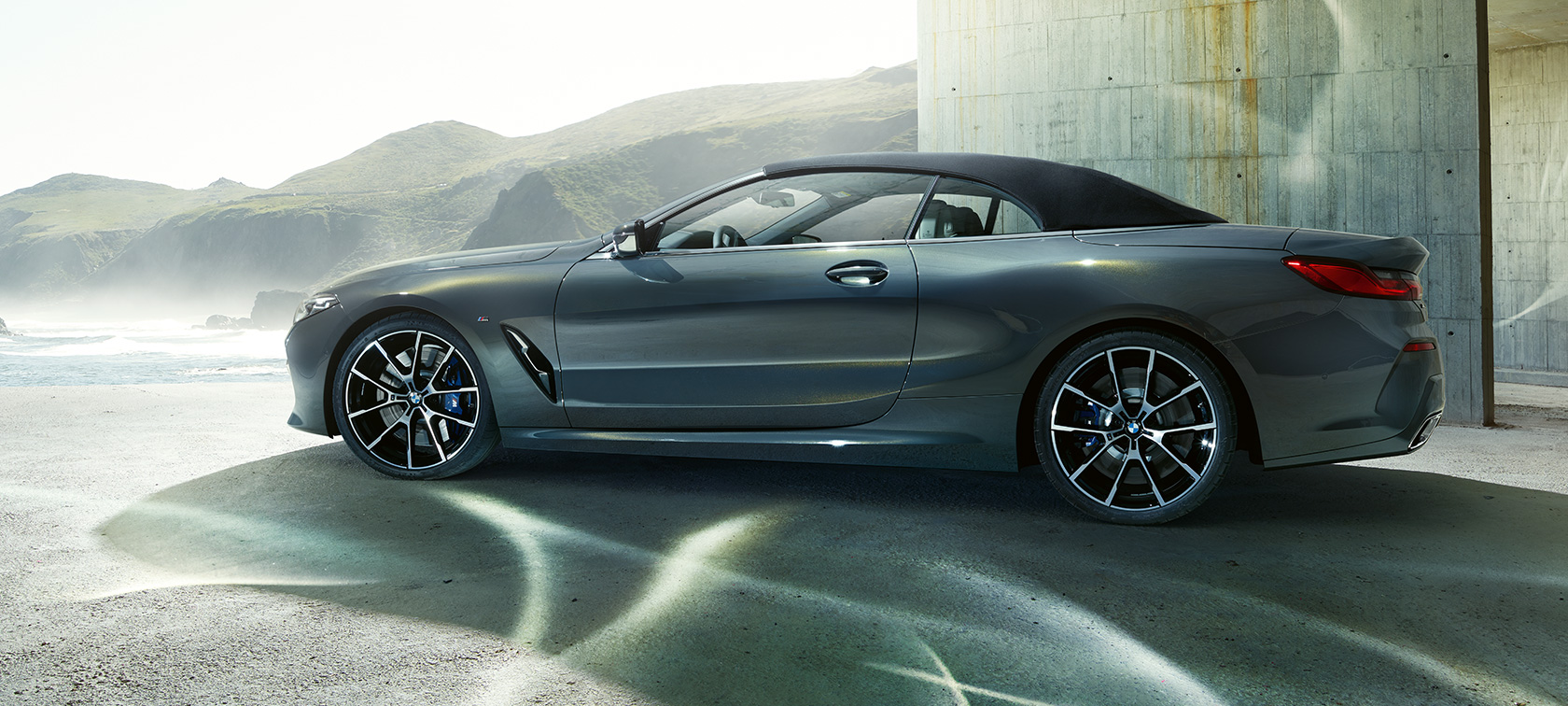 BMW M850i xDrive Cabrio, v profilu. BMW M8 Competition Cabrio v kovinski barvi Dravit Grey z zaprto platneno streho, v profilu pred gorami.