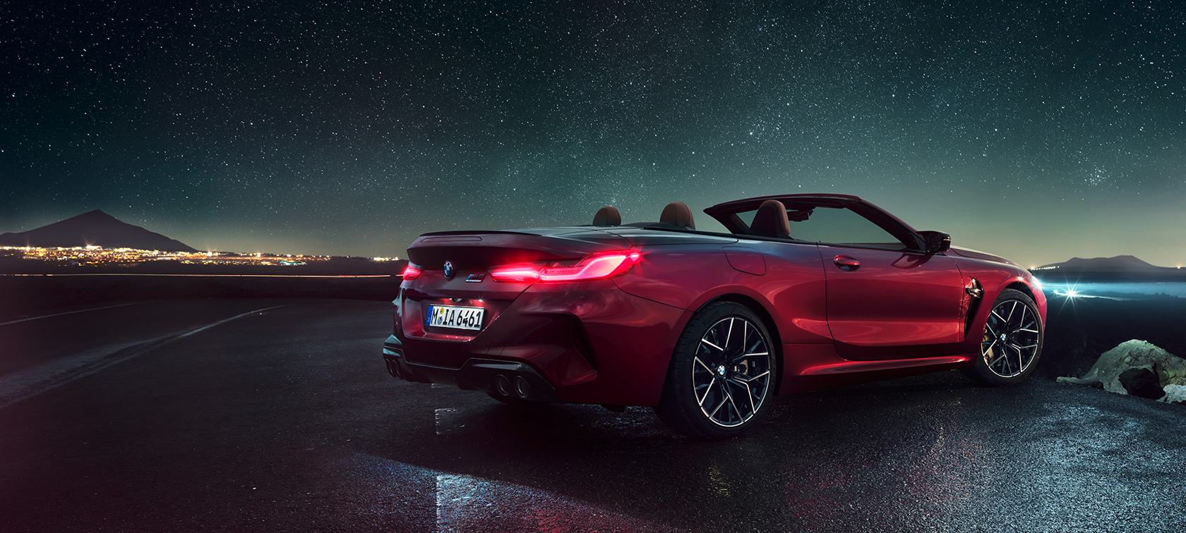 BMW M8 Competition Cabrio, v zadnjem polprofilu. BMW M8 Competition Cabrio v kovinski barvi Motegi Red z odprto platneno streho, v zadnjem polprofilu, parkiran pod zvezdnatim nebom.