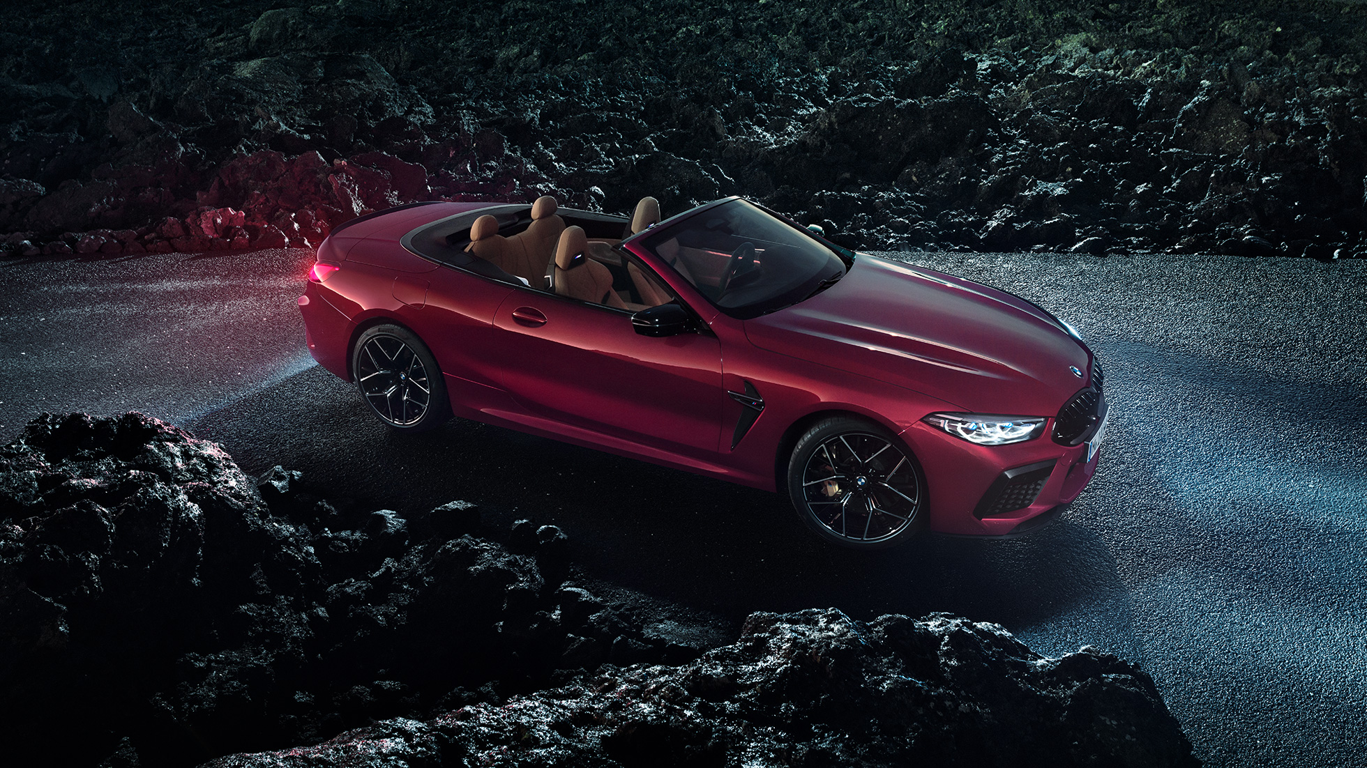 BMW M8 Competition Cabrio, v stranskem polprofilu. BMW M8 Competition Cabrio v kovinski barvi Motegi Red z odprto platneno streho, v stranskem polprofilu, parkiran na asfaltu ponoči.