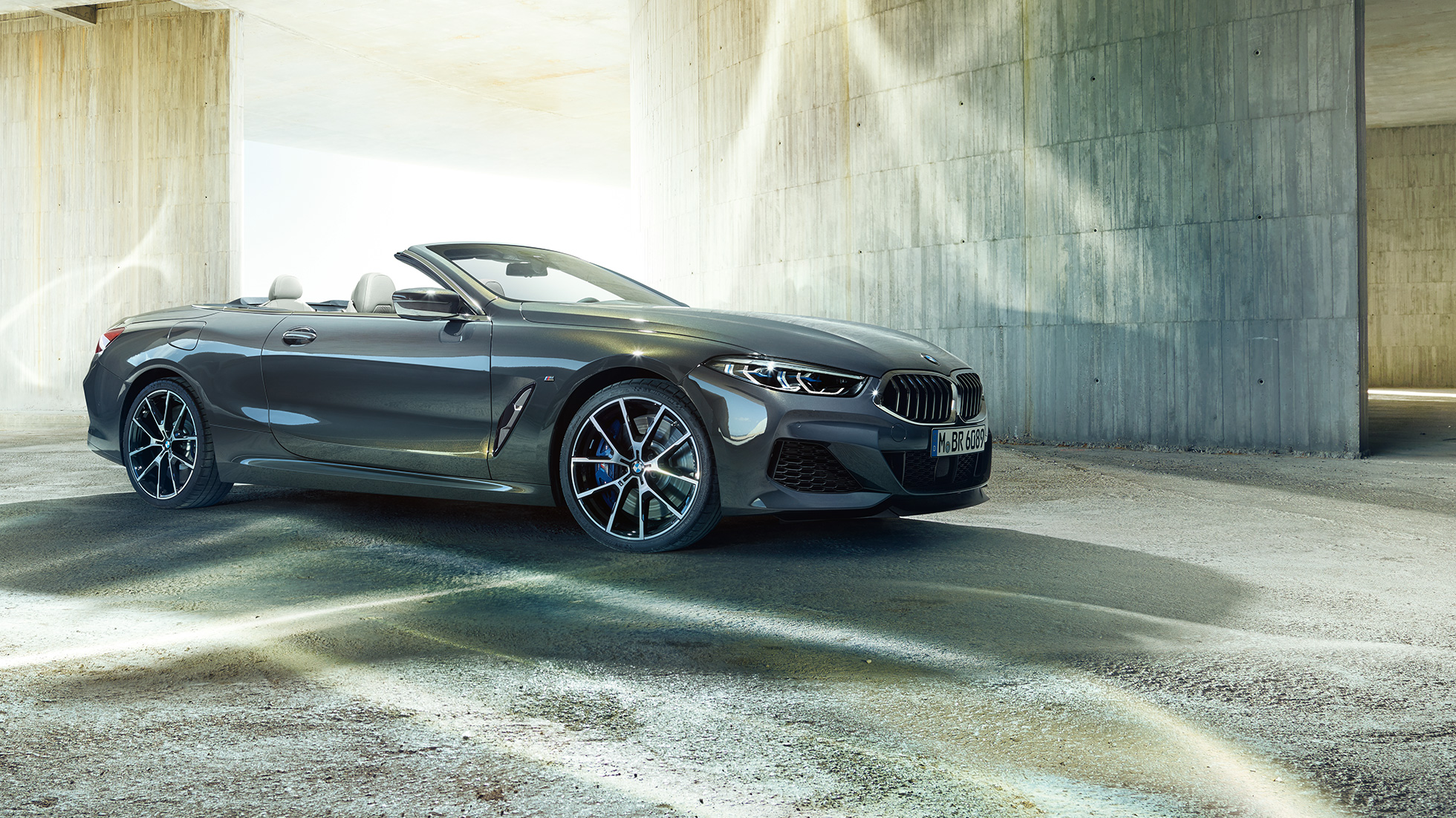 BMW M850i xDrive Cabrio, v stranskem polprofilu. BMW M850i xDrive Cabrio v kovinski barvi Dravit Grey z odprto platneno streho, v stranskem polprofilu, parkiran pred betonsko zgradbo.