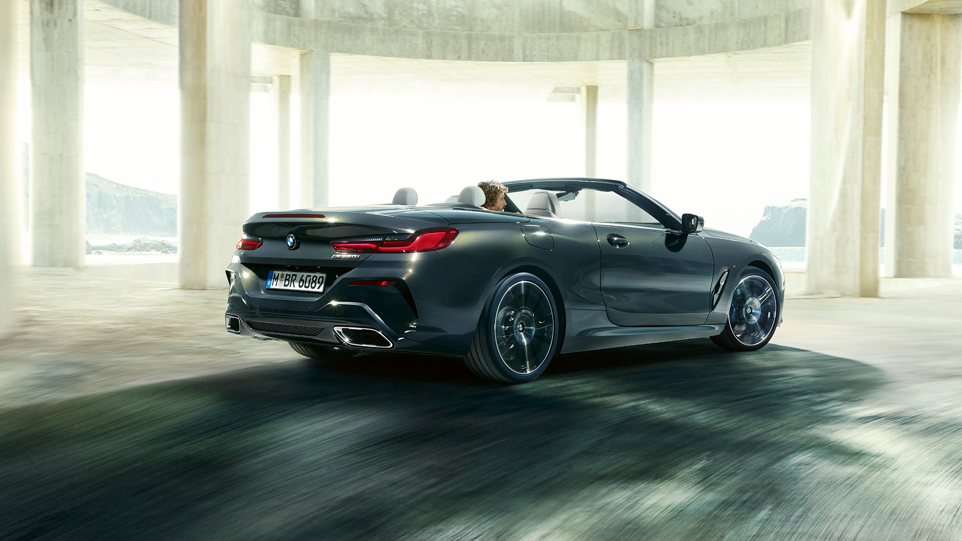 BMW M850i xDrive Cabrio, v zadnjem polprofilu. BMW M850i xDrive Cabrio v kovinski barvi Dravit Grey z odprto platneno streho, v zadnjem polprofilu, med vožnjo skozi betonsko zgradbo.