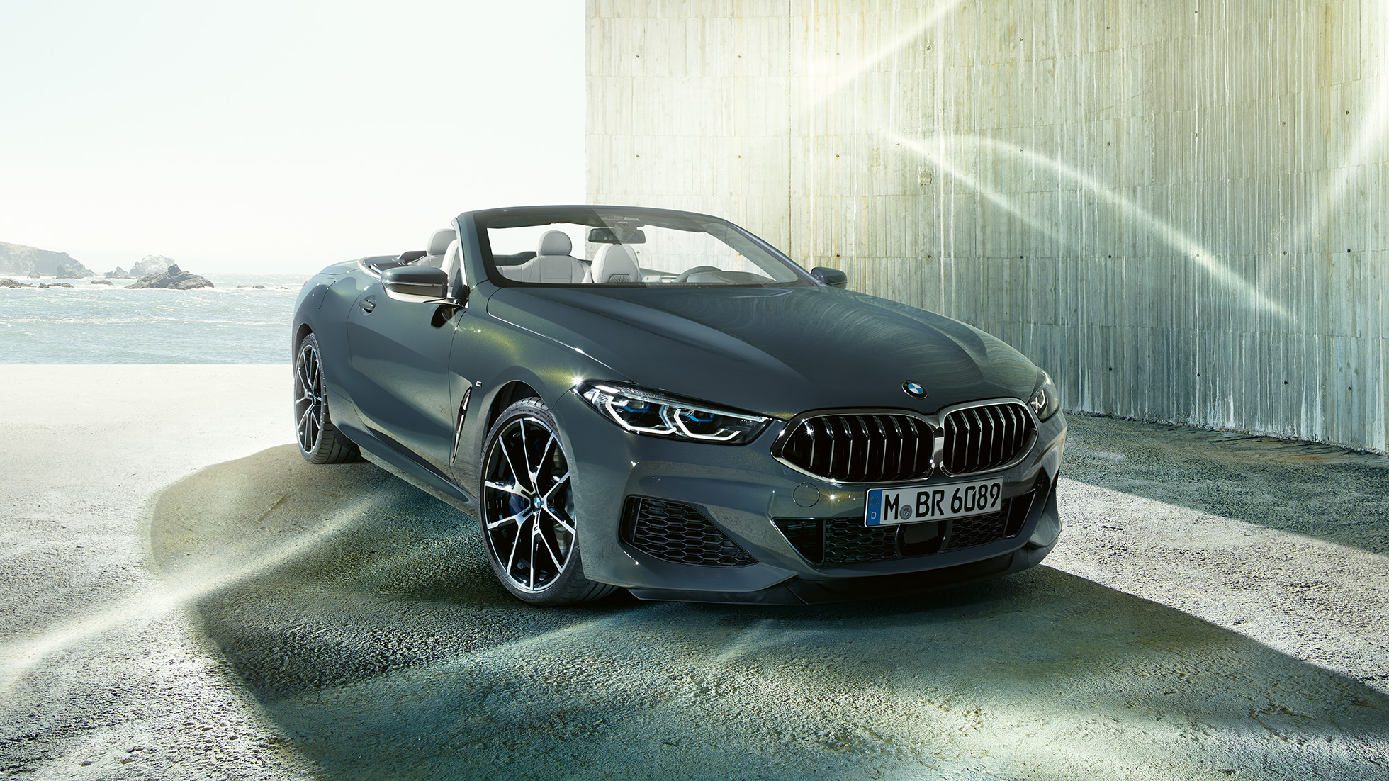 BMW M850i xDrive Cabrio, v sprednjem polprofilu. BMW M850i xDrive Cabrio v kovinski barvi Dravit Grey z odprto platneno streho, v sprednjem polprofilu, parkiran na obali.