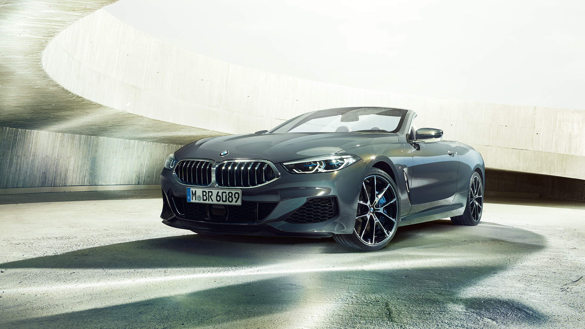 BMW M850i xDrive Cabrio, v sprednjem polprofilu. BMW M850i xDrive Cabrio v kovinski barvi Dravit Grey z odprto platneno streho, v sprednjem polprofilu, parkiran v betonski stavbi.