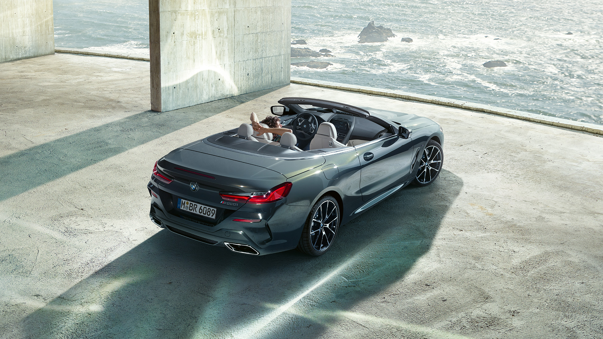 BMW M850i xDrive Cabrio, iz ptičje perspektive. BMW M850i xDrive Cabrio v kovinski barvi Dravit Grey z odprto platneno streho, iz ptičje perspektive, parkiran v betonski stavbi pred morjem.