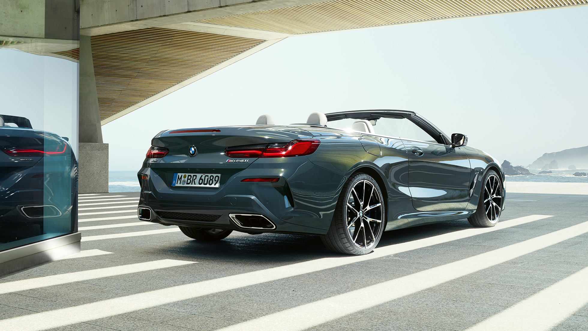 BMW M850i xDrive Cabrio, v zadnjem polprofilu. BMW M850i xDrive Cabrio v kovinski barvi Dravit Grey z odprto platneno streho, v zadnjem polprofilu, parkiran pred morjem.