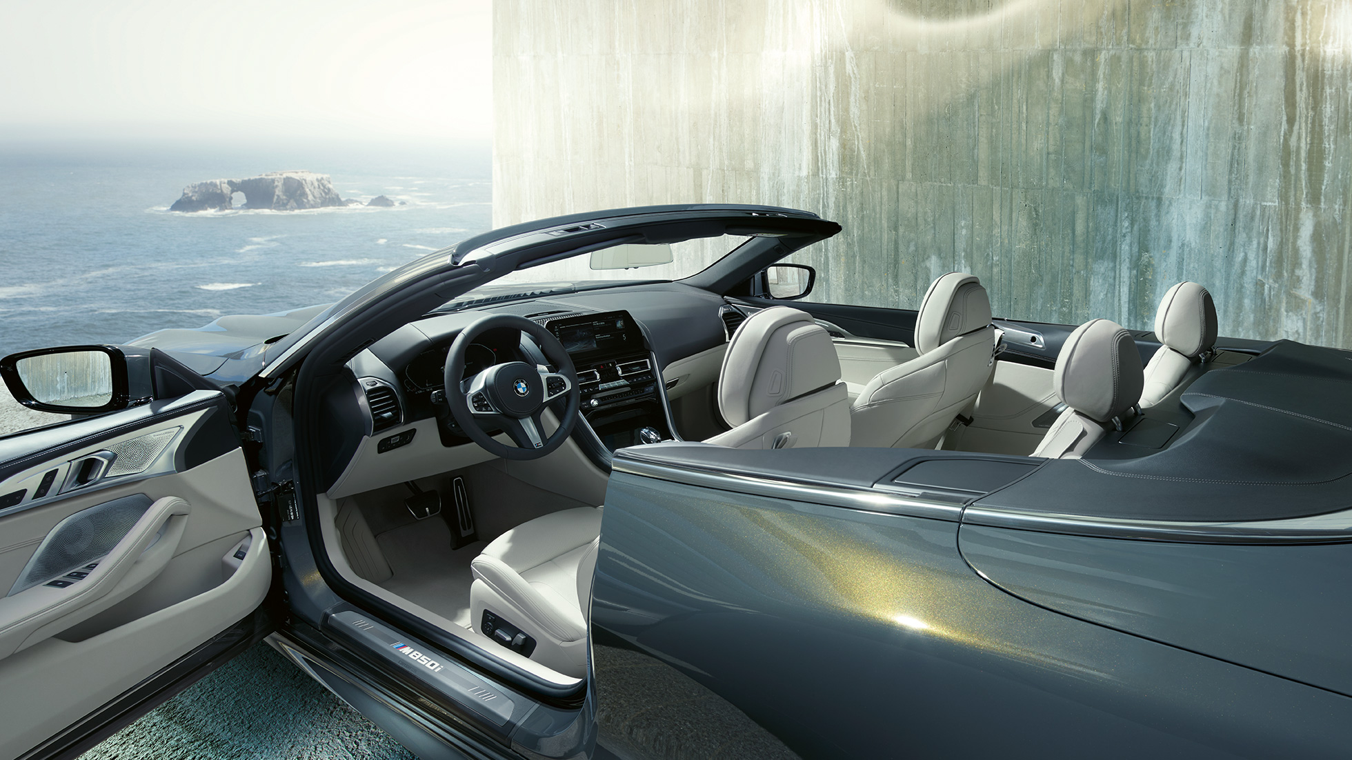BMW M850i xDrive Cabrio, notranjost. BMW M850i xDrive Cabrio v kovinski barvi Dravit Grey z odprto platneno streho, notranjost z usnjenim volanom M in obrobe BMW Individual iz usnja »Merino« v barvi Ivory White.