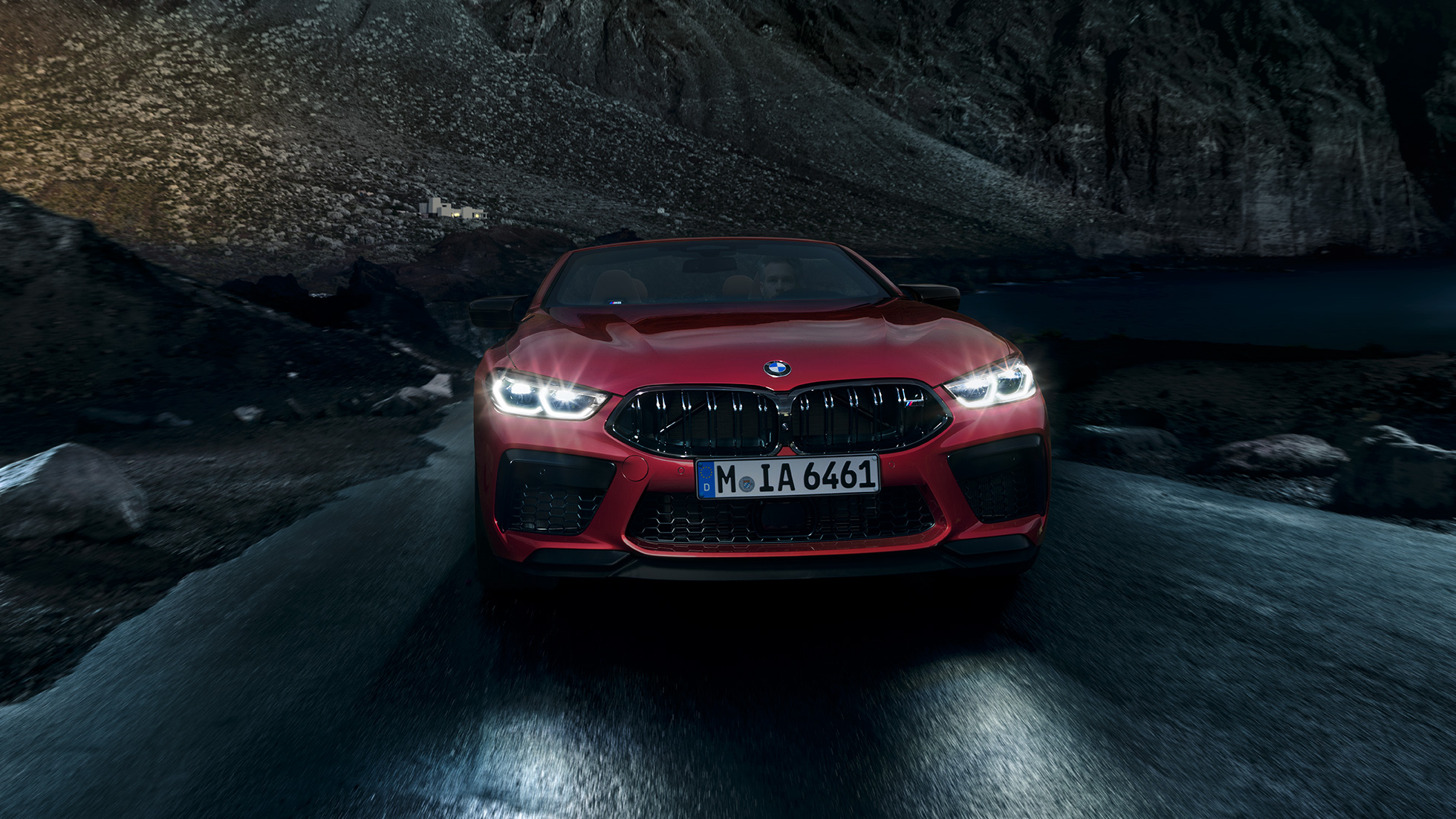 BMW M8 Competition Cabrio, s sprednje strani. BMW M8 Competition Cabrio v kovinski barvi Motegi Red z bleščečimi žarometi, s sprednje strani, med nočno vožnjo.