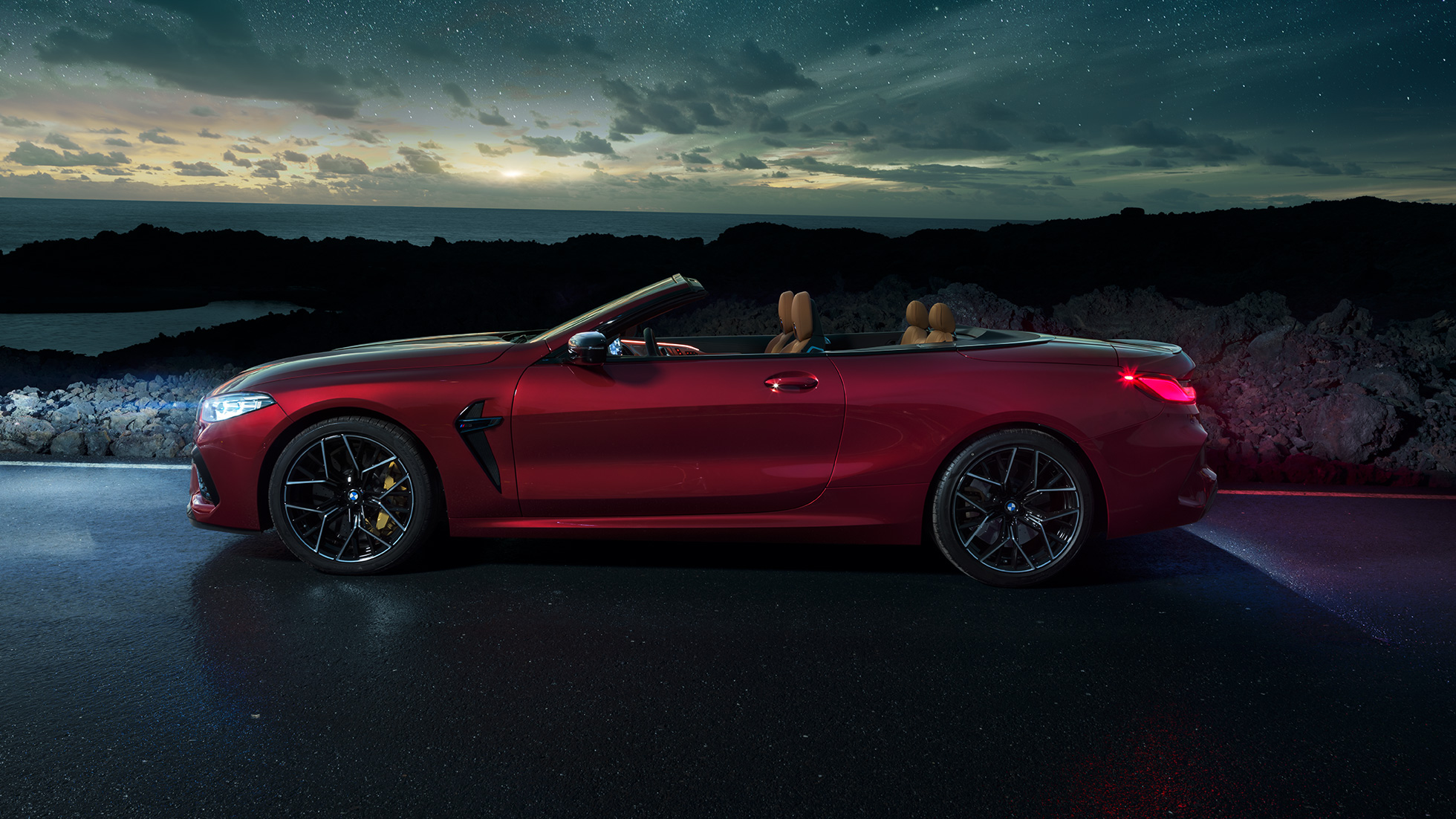 BMW M8 Competition Cabrio, v profilu. BMW M8 Competition Cabrio v kovinski barvi Motegi Red z odprto platneno streho, v profilu, parkiran pod zvezdnatim nebom.