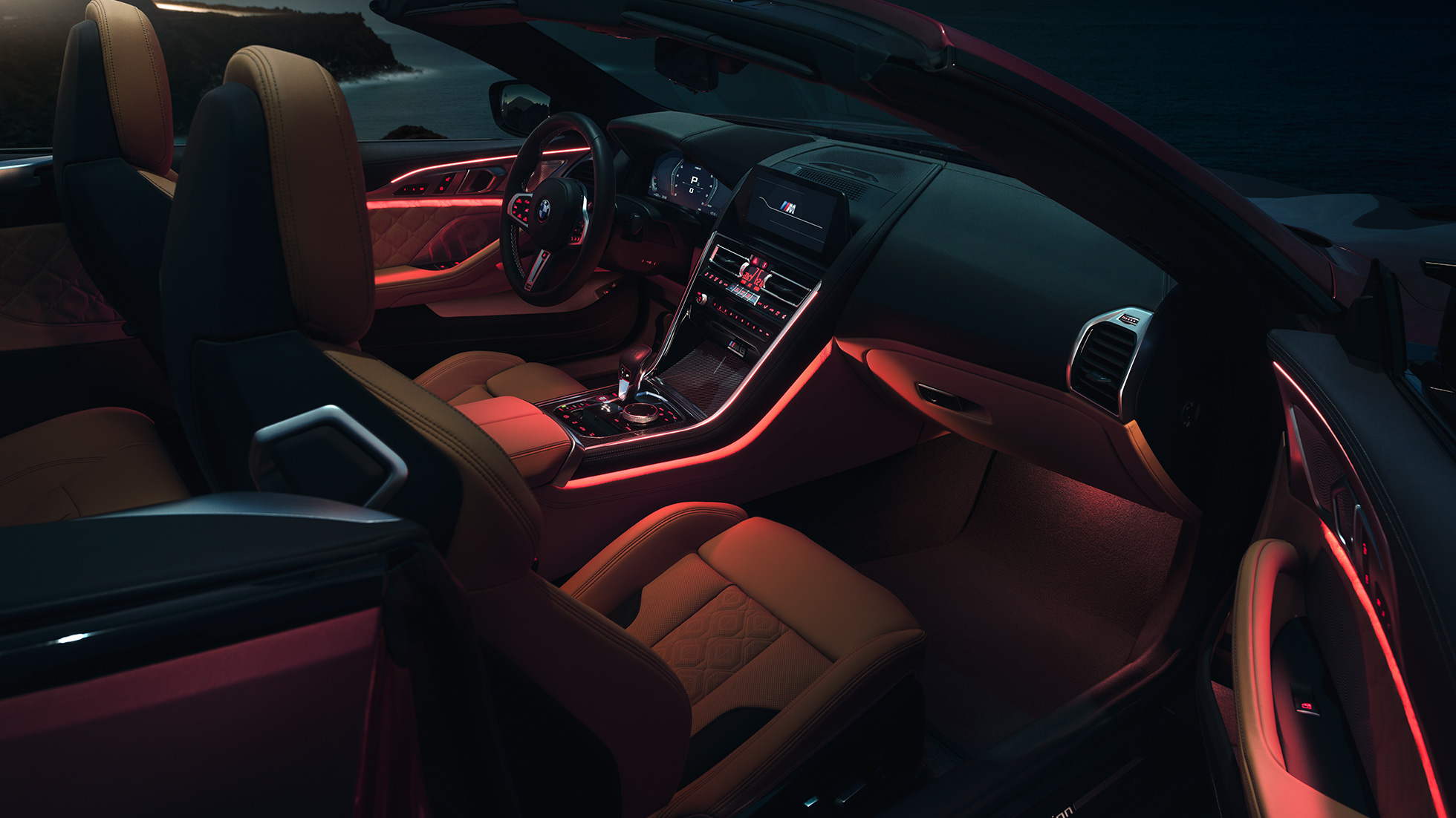 BMW M8 Competition Cabrio, notranjost. BMW M8 Competition Cabrio z odprto platneno streho, notranjost z rdečo ambientalno osvetlitvijo ponoči.