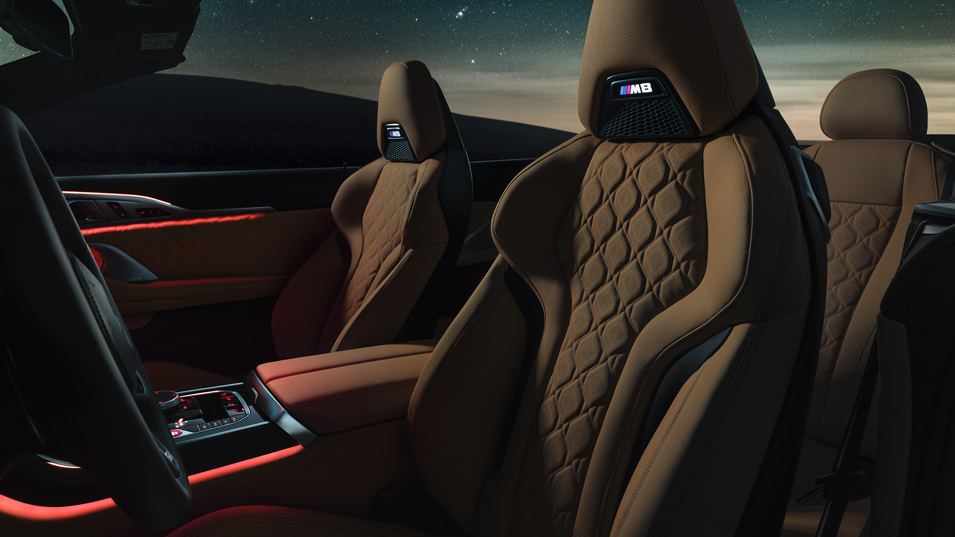 M športni sedeži v usnju »Merino«. BMW M8 Competition Cabrio z odprto platneno streho pod zvezdnatim nebom, notranjost z M športnimi sedeži v usnju »Merino« in rdečo ambientalno svetlobo.