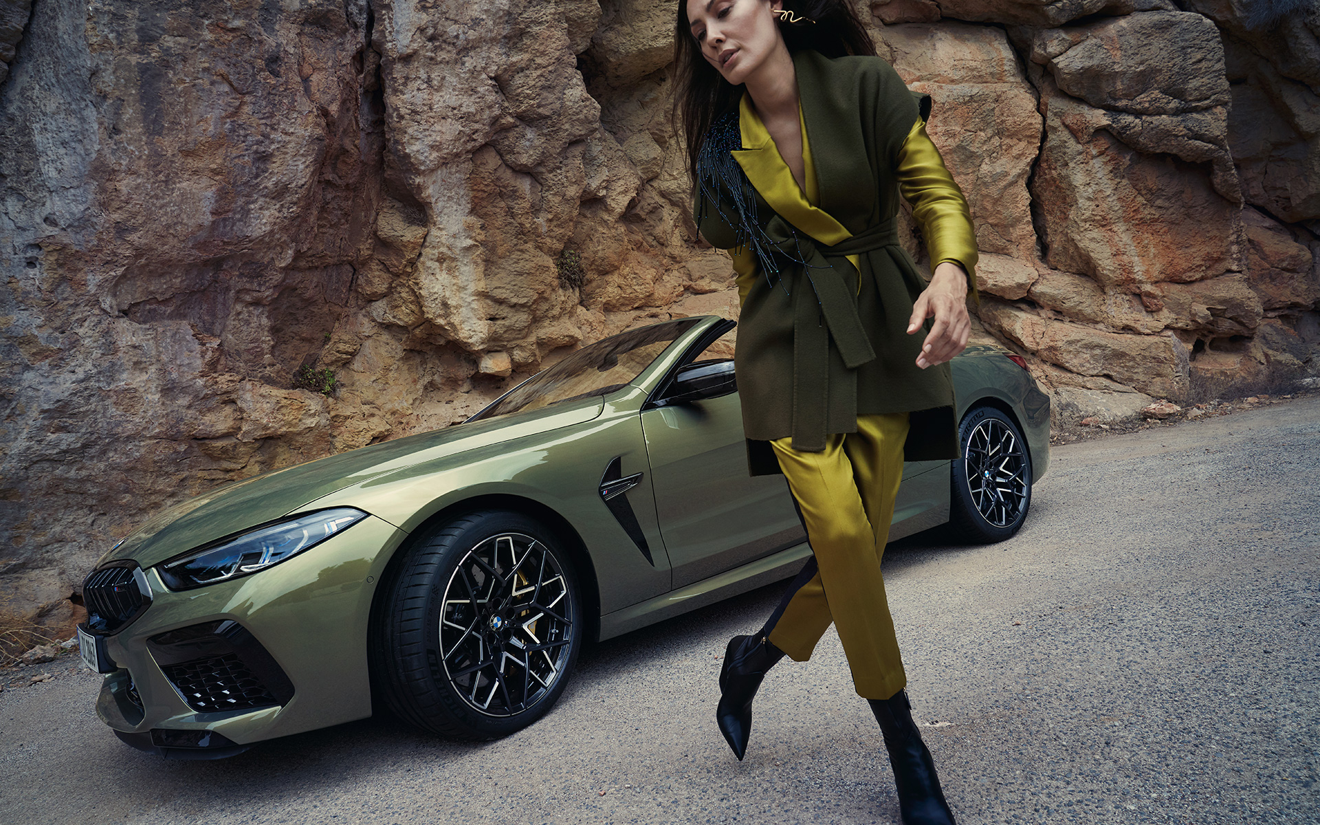 BMW M8 Competition Cabrio, F91, LCI, 2022, tričetrtinski pogled od spredaj med mirovanjem na kamnitem terenu BMW M8 Competition Cabrio, F91, LCI, osvežitev 2022, lakiranje BMW Individual v kovinski barvi Brass, tričetrtinski pogled od spredaj med mirovanjem na kamnitem terenu z manekenko