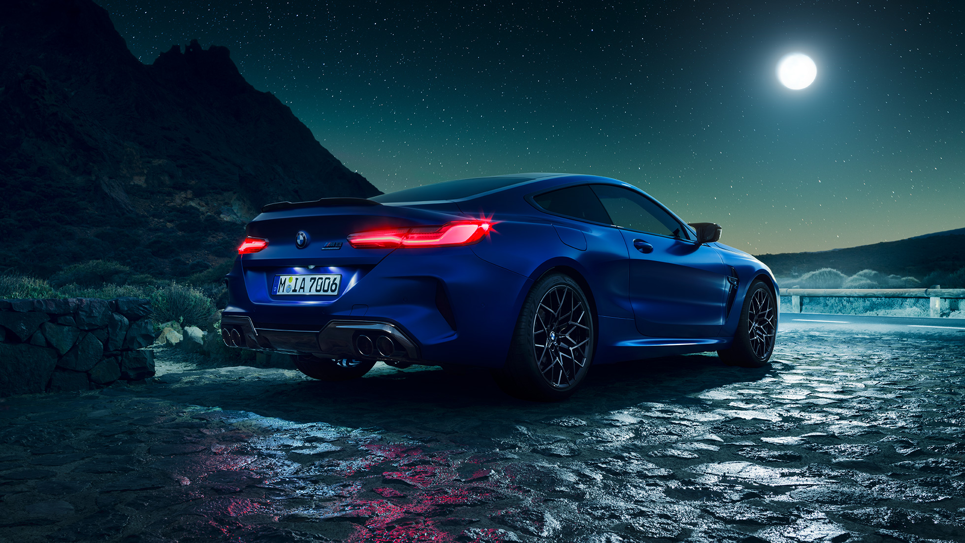 BMW M8 Competition Coupé, v polprofilu z zadnje strani. BMW M8 Competition Coupé v kovinski barvi Marina Bay Blue z osvetljenimi zadnjimi LED lučmi, v polprofilu z zadnje strani, ob ščipu.