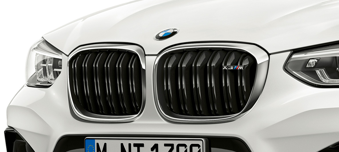 BMW X3 M, ledvičasta maska M. BMW X3 M v barvi Alpine White, zunanjost, sprednji del, ledvičasta maska M s kromiranimi poudarki in oznako modela.