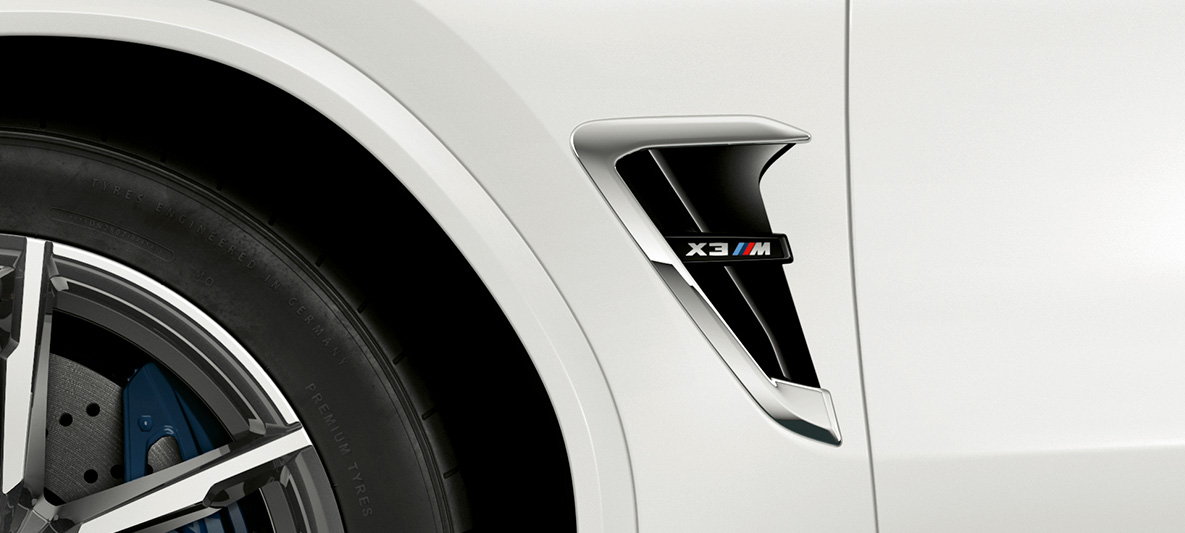 BMW X3 M, maska M. BMW X3 M v barvi Alpine White, zunanjost, bočna maska M s kromiranimi poudarki in oznako modela.