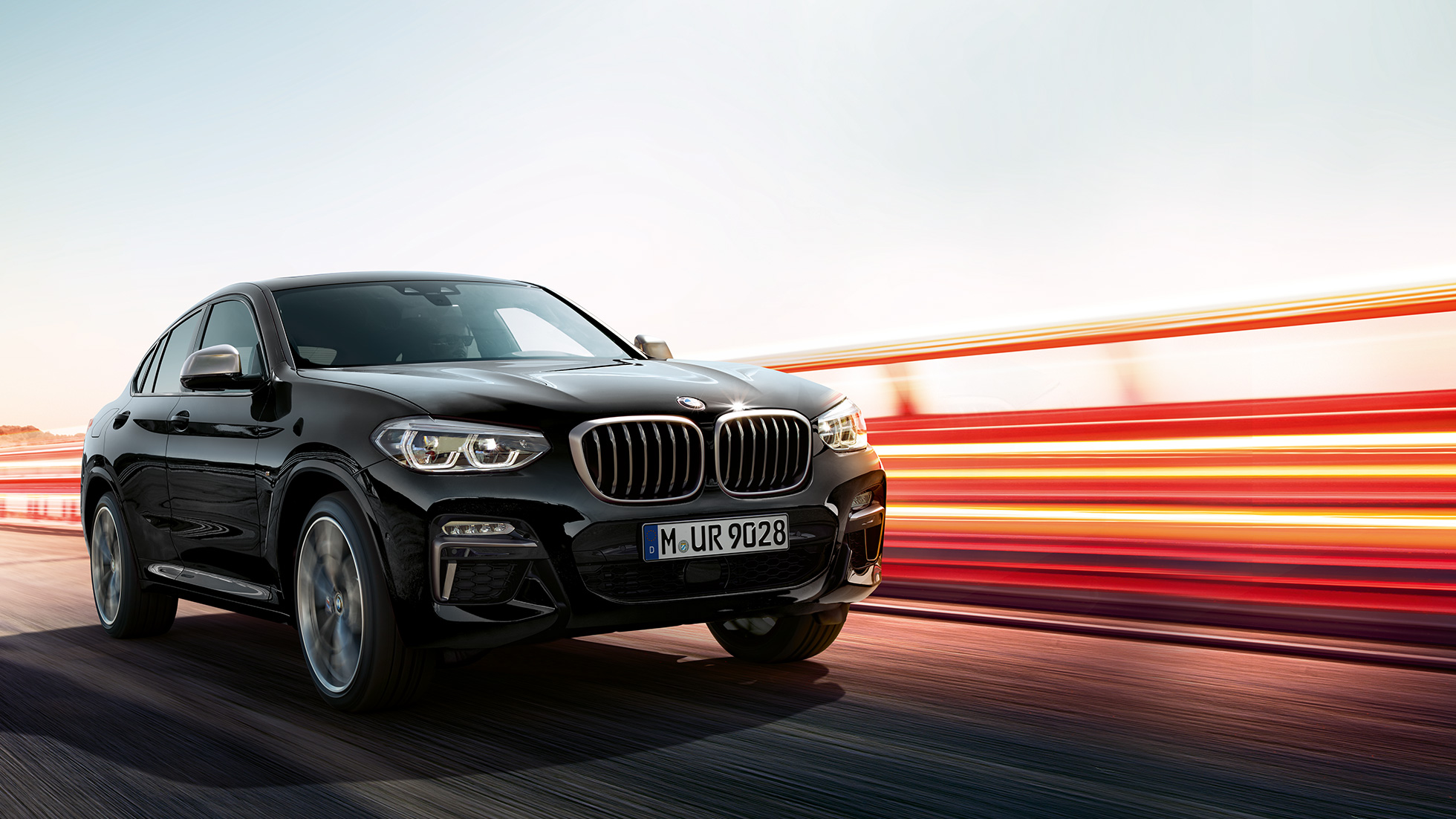 BMW X4 M40i, v polprofilu z zadnje strani. BMW X4 M40i v kovinski barvi Black Sapphire, med vožnjo, zunanjost, v polprofilu od zadaj s svetlobnimi črtami.