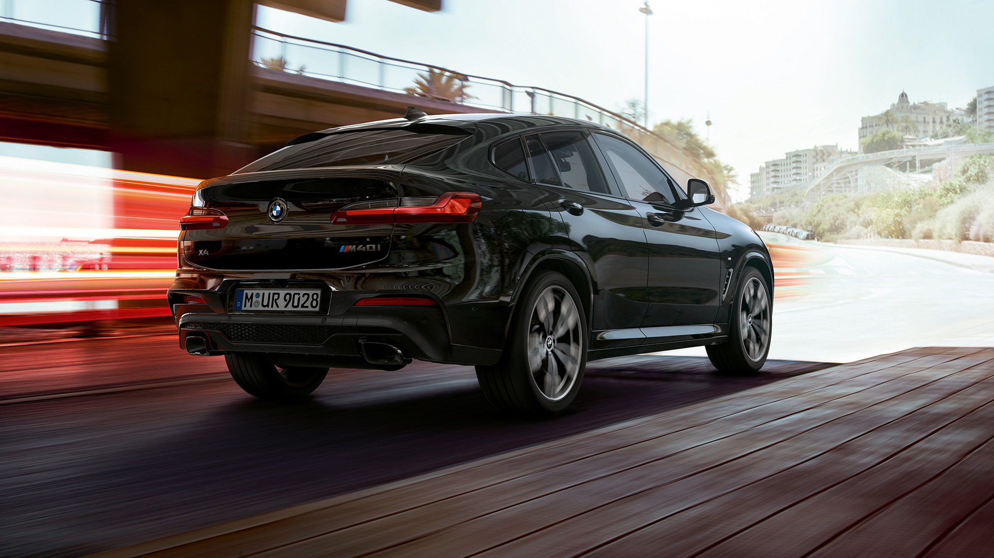 BMW X4 M40i, v polprofilu z zadnje strani. BMW X4 M40i v kovinski barvi Black Sapphire, zunanjost, v polprofilu od zadaj, od strani s svetlobnimi sledmi.