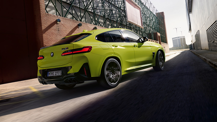 BMW X4 M Competition, F98, LCI, 2021, tričetrtinski pogled od spredaj BMW X4 M Competition, F98, LCI, osvežitev 2021, barva Sao Paulo Yellow, tričetrtinski pogled od spredaj pri vožnji med tovarniškimi zgradbami