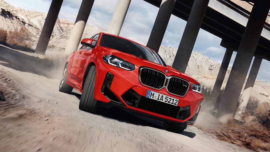 BMW X4 M, F98, LCI, osvežitev 2021, kovinska barva Toronto Red, tričetrtinski pogled od spredaj pred stebri pod mostom