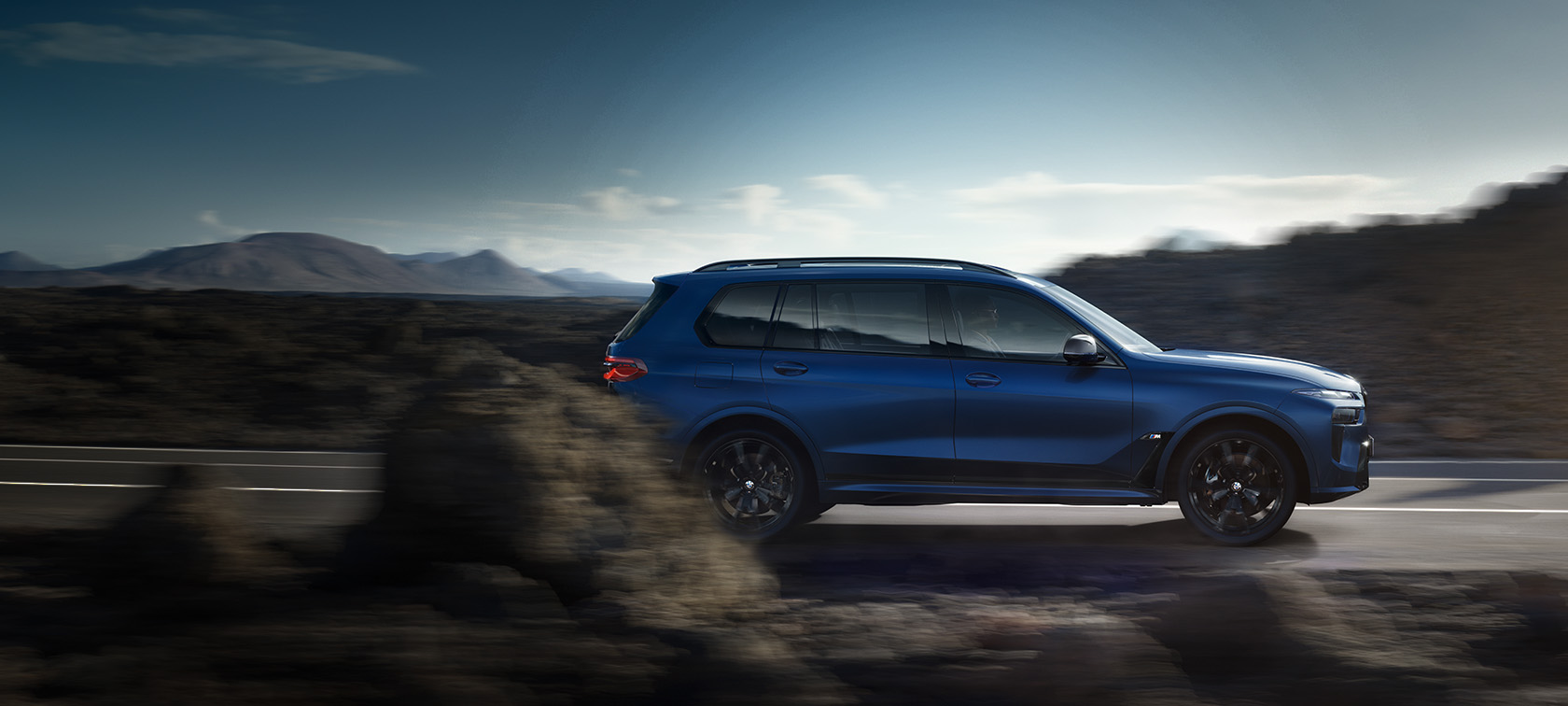 BMW X7 M60i xDrive G07 LCI Frozen Marina Bay Modri stranski pogled na vožnjo po ulici BMW X7 M60i xDrive G07 LCI Frozen Marina Bay Modri stranski pogled na vožnjo po ulici pred gorsko pokrajino