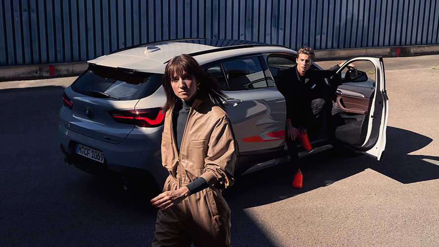 BMW X2, F39, barva Brooklyn Grey, tričetrtinski pogled od zadaj z odprtimi vrati in dvema osebama BMW X2, F39, barva Brooklyn Grey, tričetrtinski pogled od zadaj z odprtimi vrati in dvema osebama