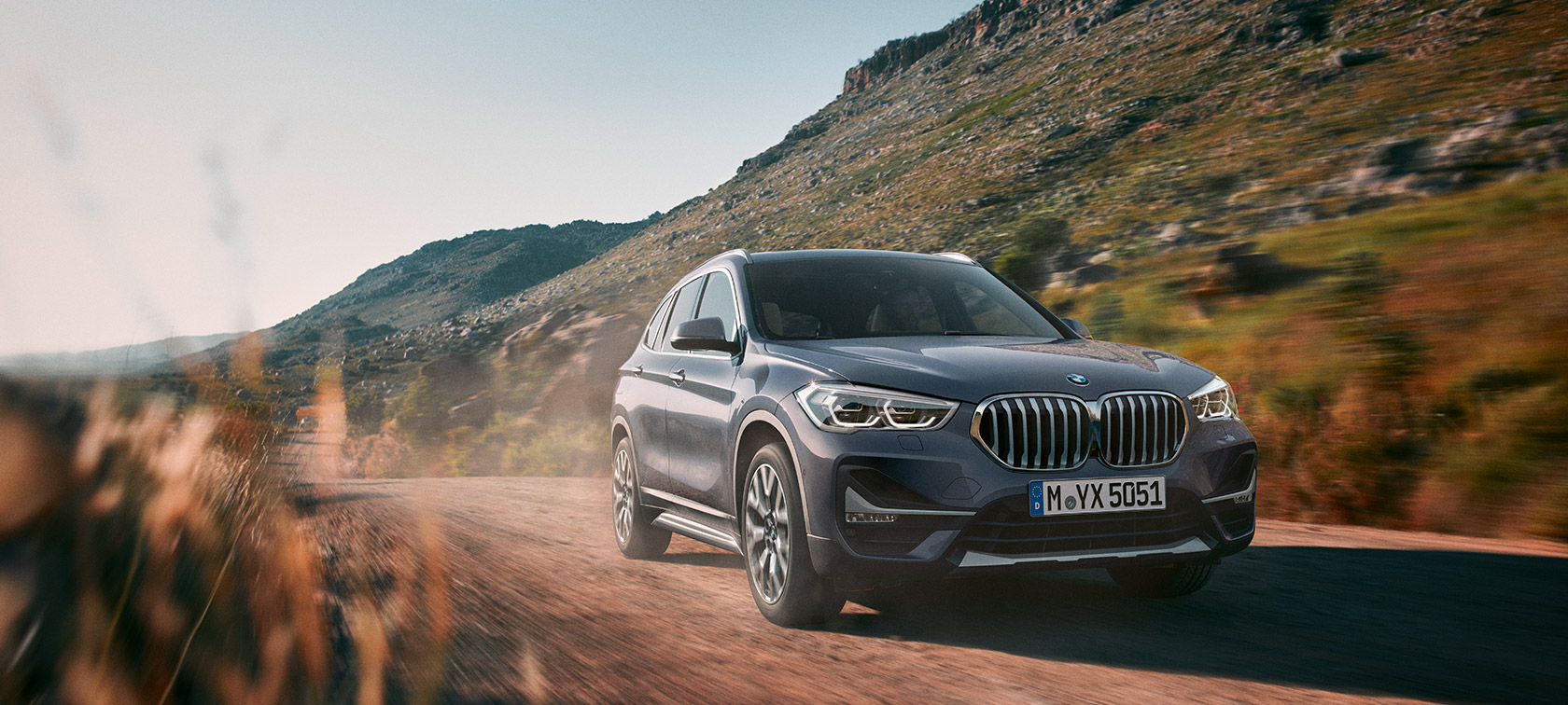 BMW X1, tričetrtinski pogled od spredaj med vožnjo pred gorsko pokrajino.