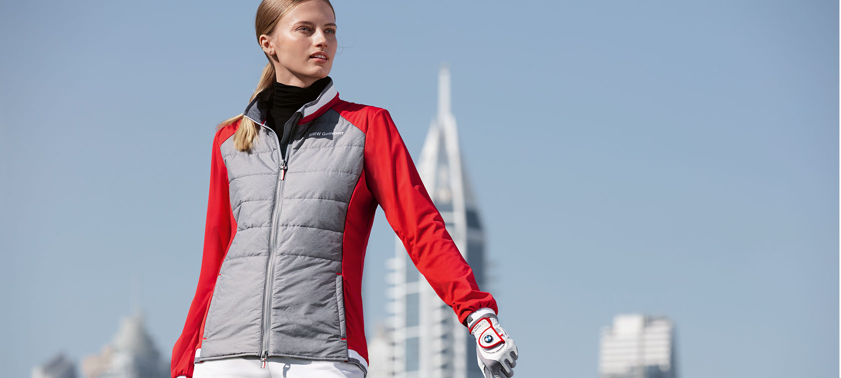 Ženska jakna iz kolekcije BMW Golfsport. Mlada ženska na sliki predstavlja vrhunske izdelke iz kolekcije BMW Golfsport, v ozadju pa vidimo obzorje velikega mesta.