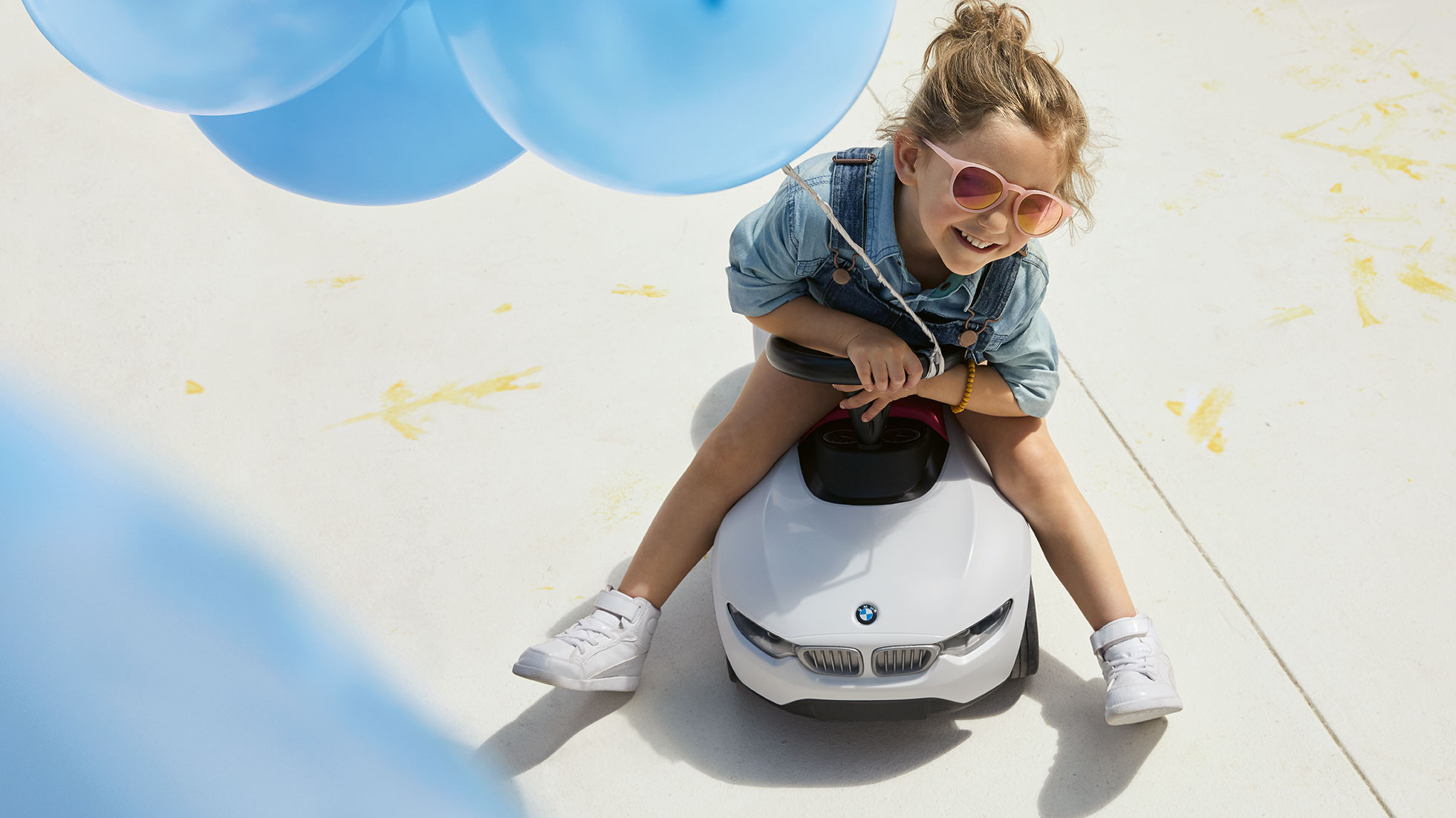 Kolekcija BMW Kids. Deklica z roza sončnimi očali in modrim balonom zadovoljno sedi na otroškem poganjalcu BMW Baby Racer.