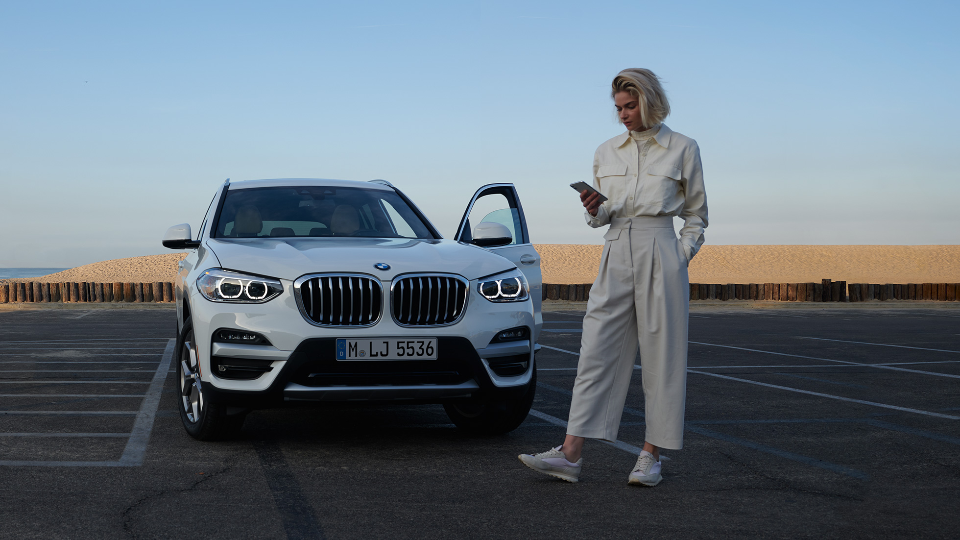 Ženska uporablja aplikacijo My BMW na pametnem telefonu Ženska uporablja aplikacijo My BMW iz BMW ConnectedDrive na pametnem telefonu pred BMW X5 bele barve