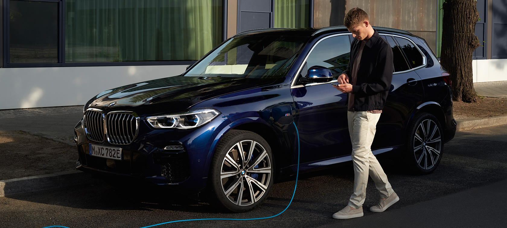Človek uporablja aplikacijo My BMW na pametnem telefonu Moški uporablja aplikacijo My BMW iz BMW ConnectedDrive na pametnem telefonu, medtem ko polni svoj priključni hibrid BMW X5